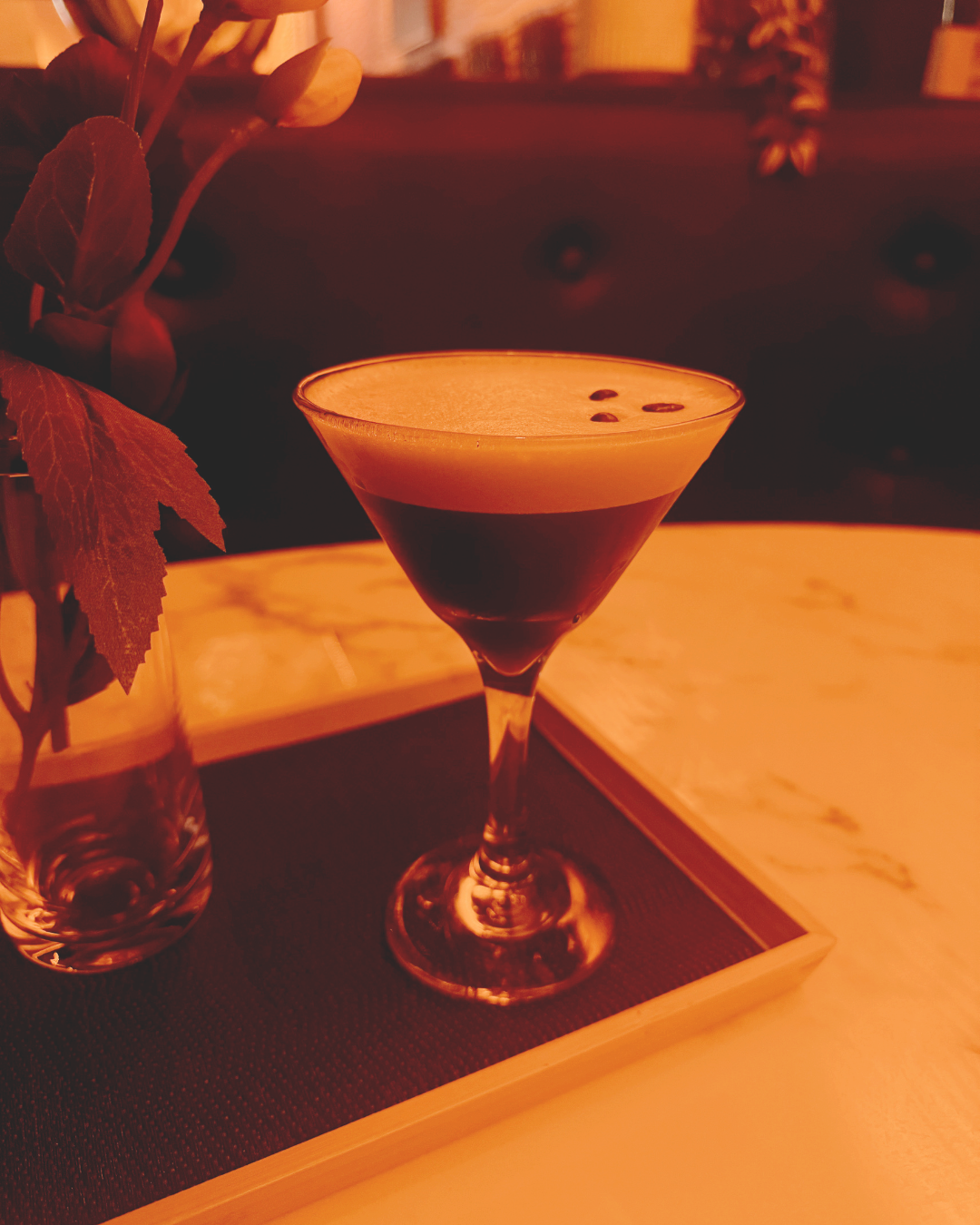 A cocktail in a martini glass with a creamy beige top and dark liquid base, garnished with a few small dark seeds, on a table next to a magazine and a vase with leaves.