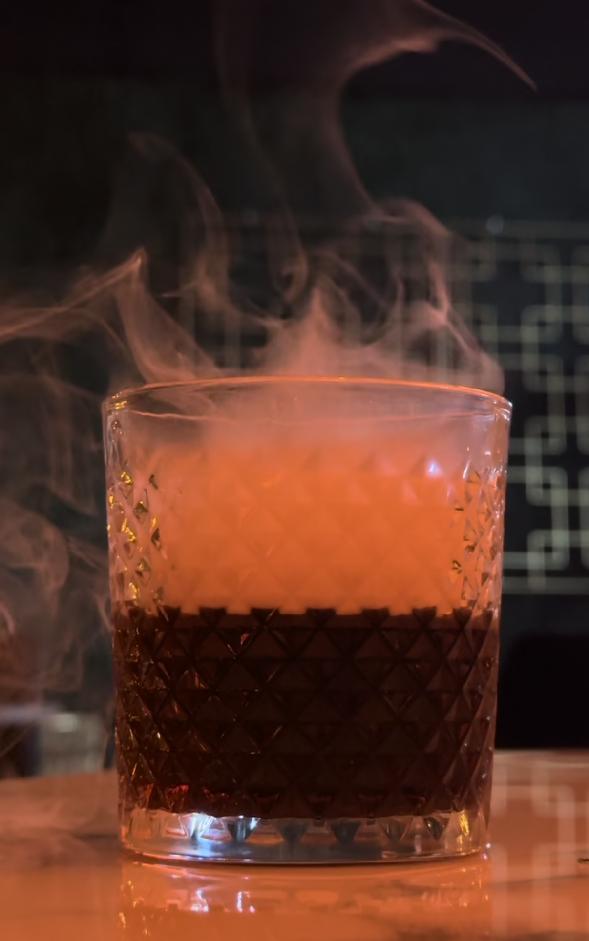 A close-up of a glass with a geometric pattern, filled with an orange-colored beverage. Steam or vapor rises from the drink.