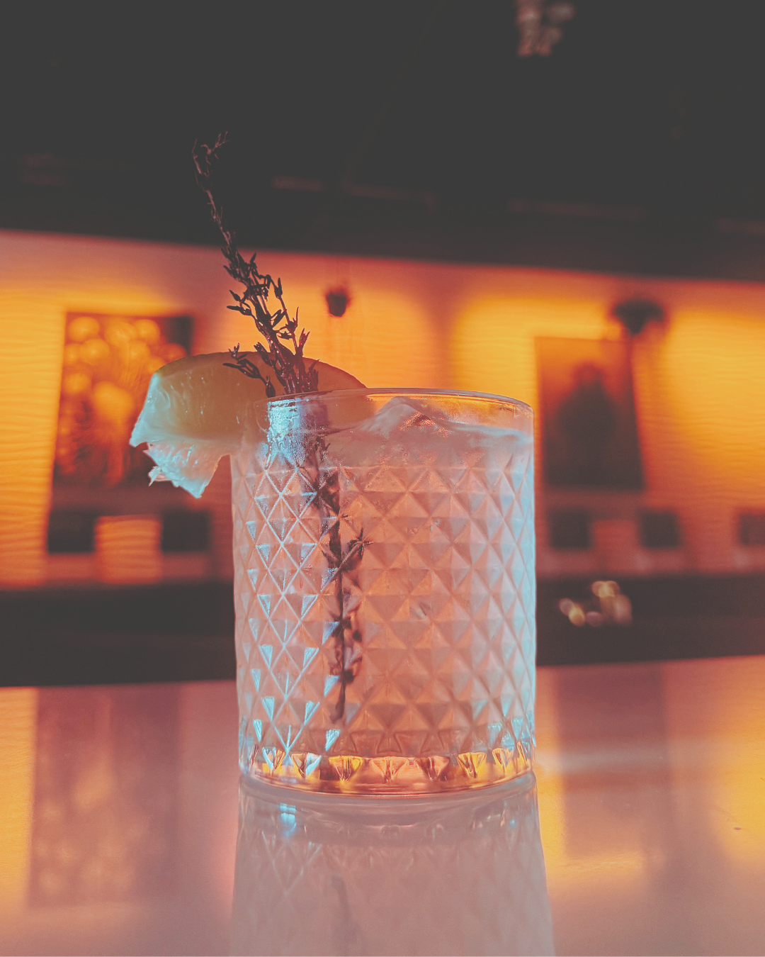 A glass with a textured diamond pattern contains a pink cocktail, garnished with a lemon wedge and a sprig of herbs, on a reflective surface in a dimly lit bar with warm orange lighting.