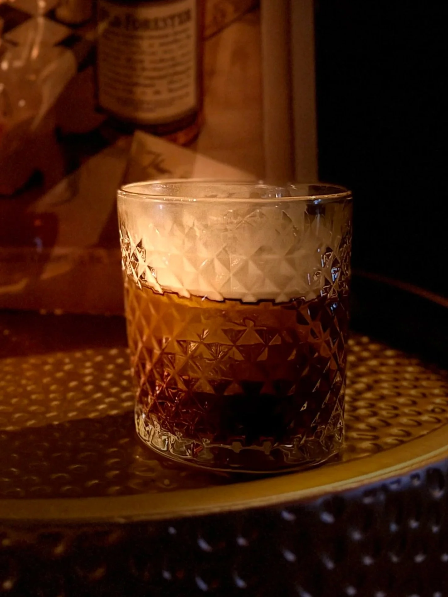 A glass of amber-colored beverage on a wooden table, illuminated by a candle inside a decorative glass holder, with a blurred background.