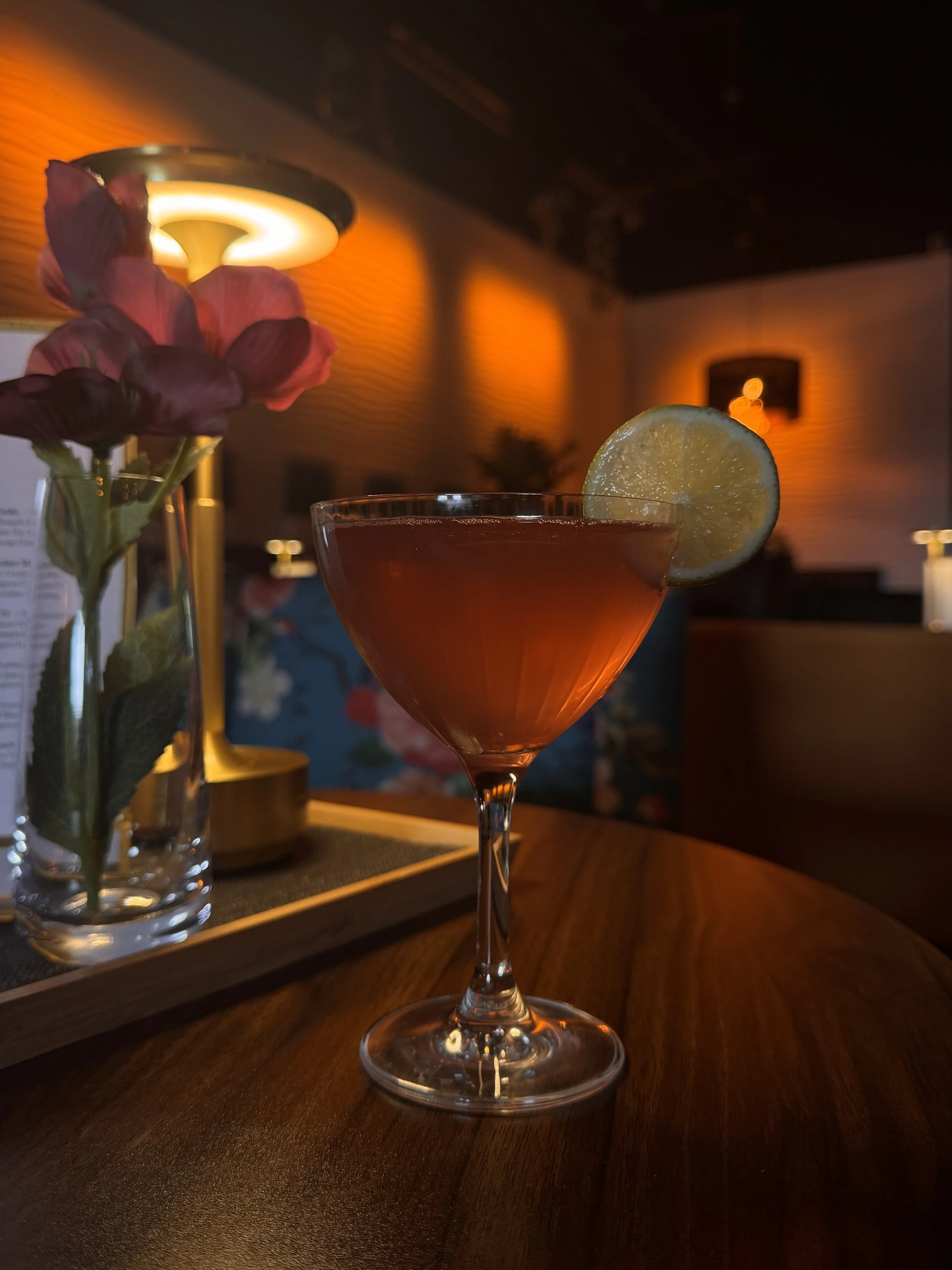 A cocktail glass with a lemon wedge garnish on the rim, placed on a wooden table in a dimly lit bar or restaurant setting.