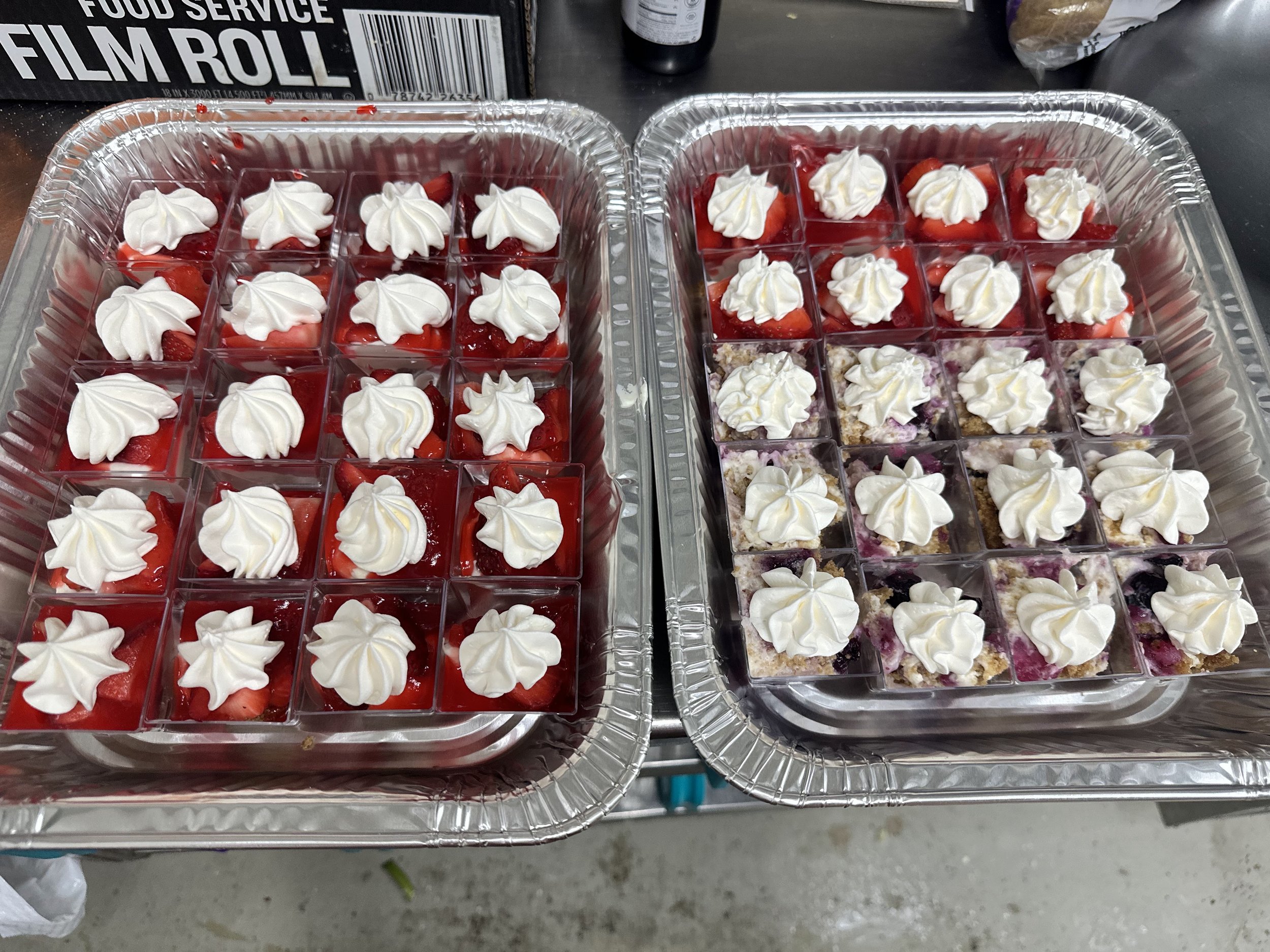 Two aluminum trays filled with individual square servings of dessert topped with whipped cream, one with strawberries and the other with mixed berries.