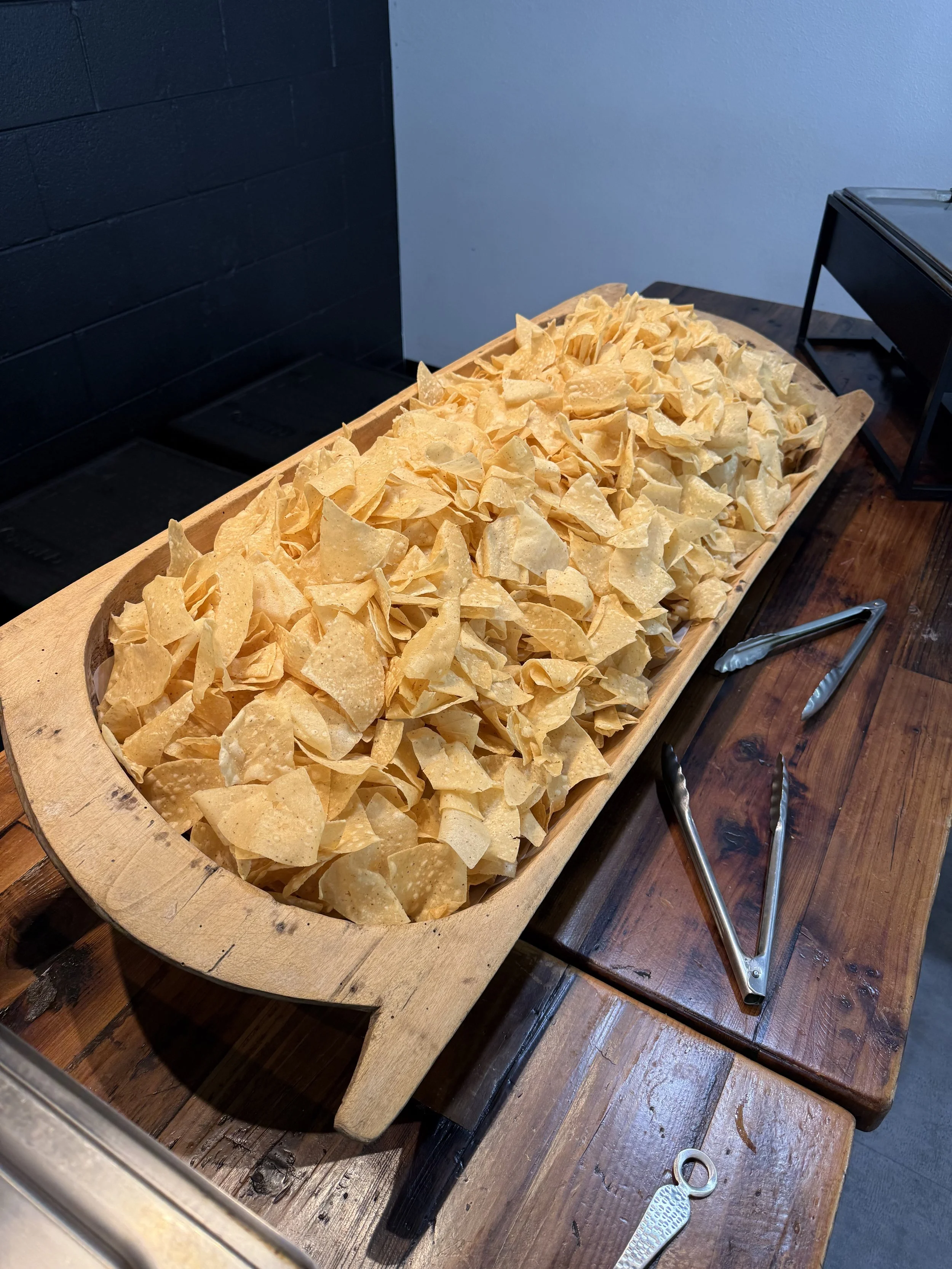 A large wooden tray filled with potato chips on a wooden table, with tongs and a chip clip nearby, against a background of a black wall and a light blue wall.