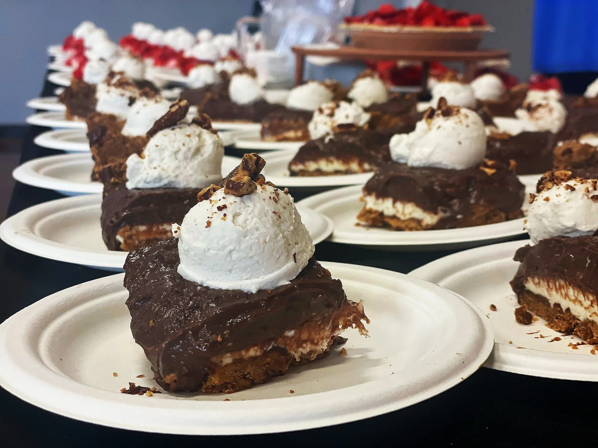 Multiple plates with dessert slices topped with whipped cream, chocolate sauce, and crushed nuts, arranged on a black surface, with some plates containing strawberries in the background.