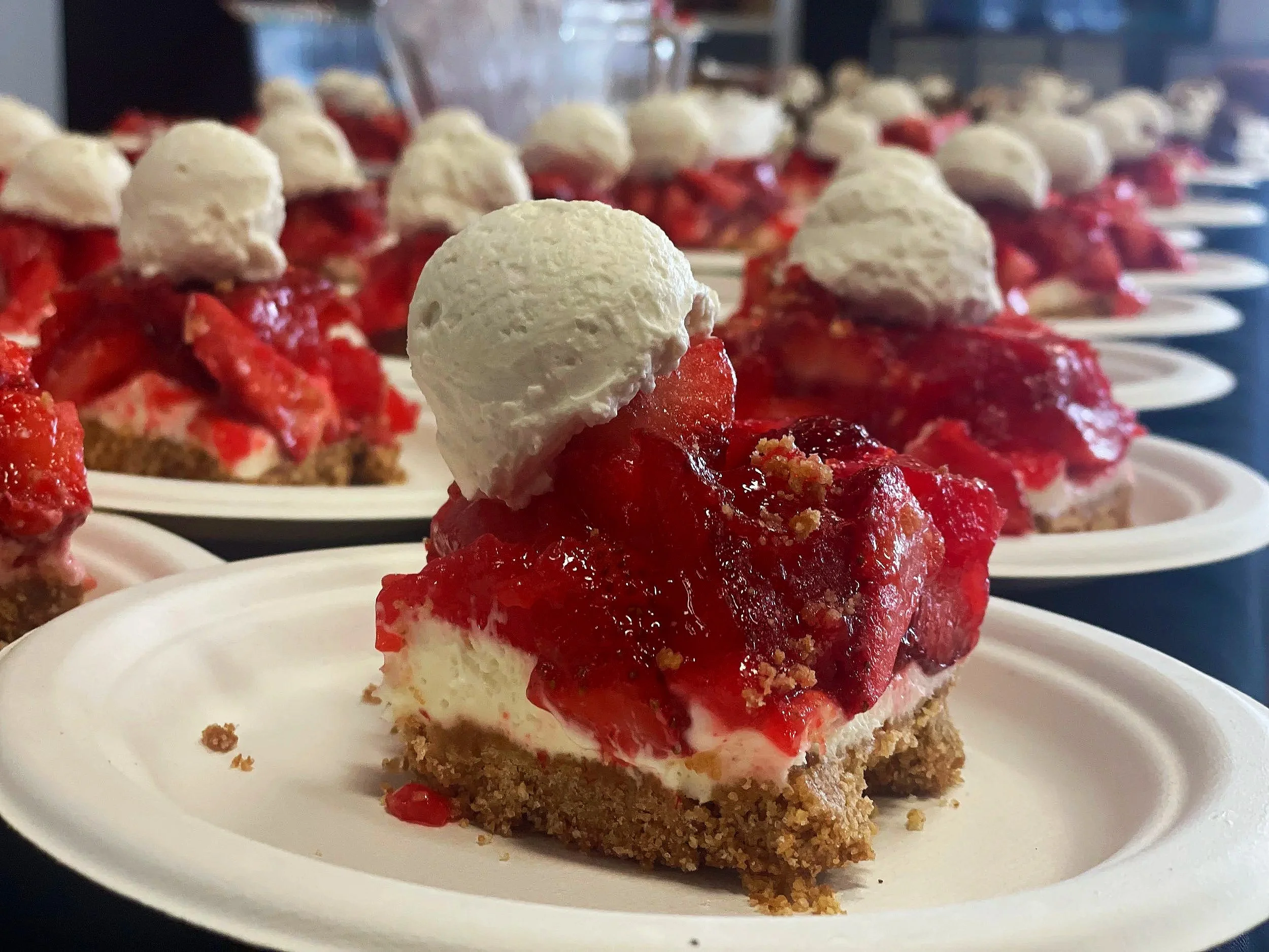 A close-up of a dessert on a white plate, consisting of a graham cracker crust, topped with a layer of red fruit (likely strawberries or cherries), a dollop of whipped cream, and additional fruit and whipped cream on top.