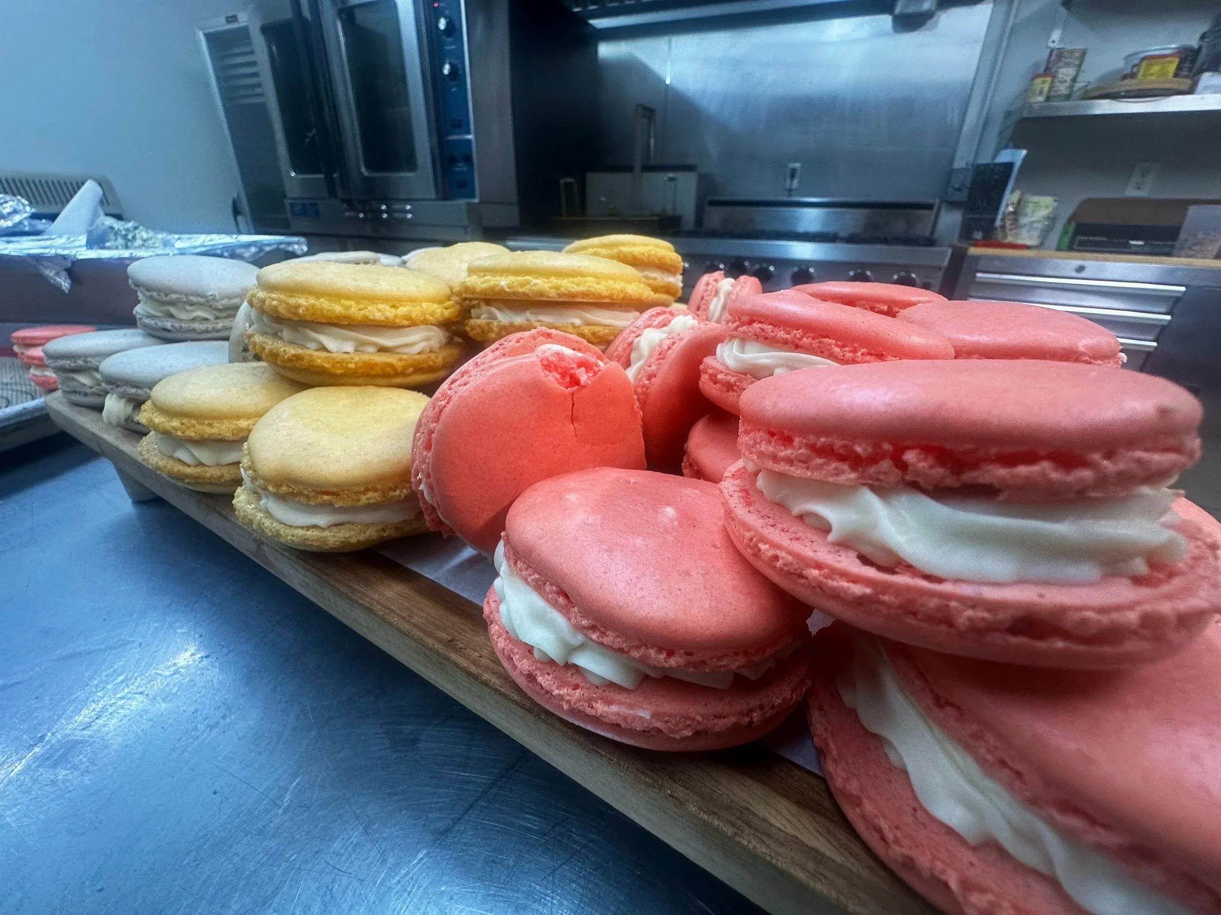 Close-up image of assorted macarons with white filling on a wooden tray. The macarons are in pastel yellow, pink, and gray colors. The background shows a commercial kitchen with stainless steel appliances and shelves.