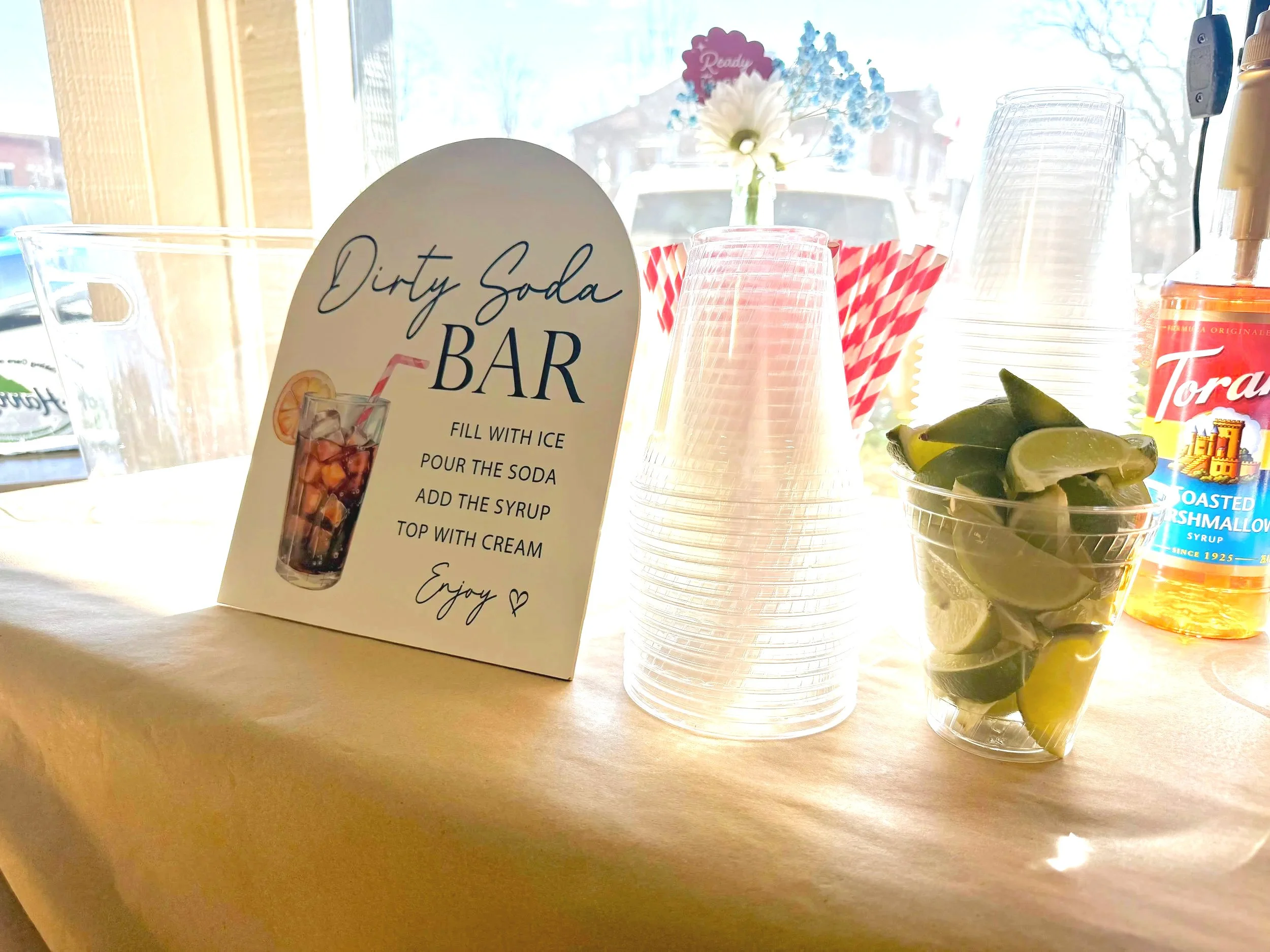 A countertop with a sign promoting a "Dirty Soda Bar," clear plastic cups, a bottle of syrup, lemon slices in a cup, and a jar with flowers, with sunlight coming through a window.