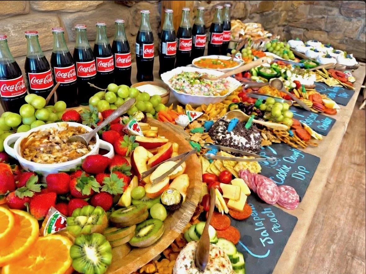 A buffet table with a variety of food including fresh fruits, salads, cold beverages in glass bottles, and assorted snacks and desserts, set against a stone wall backdrop.