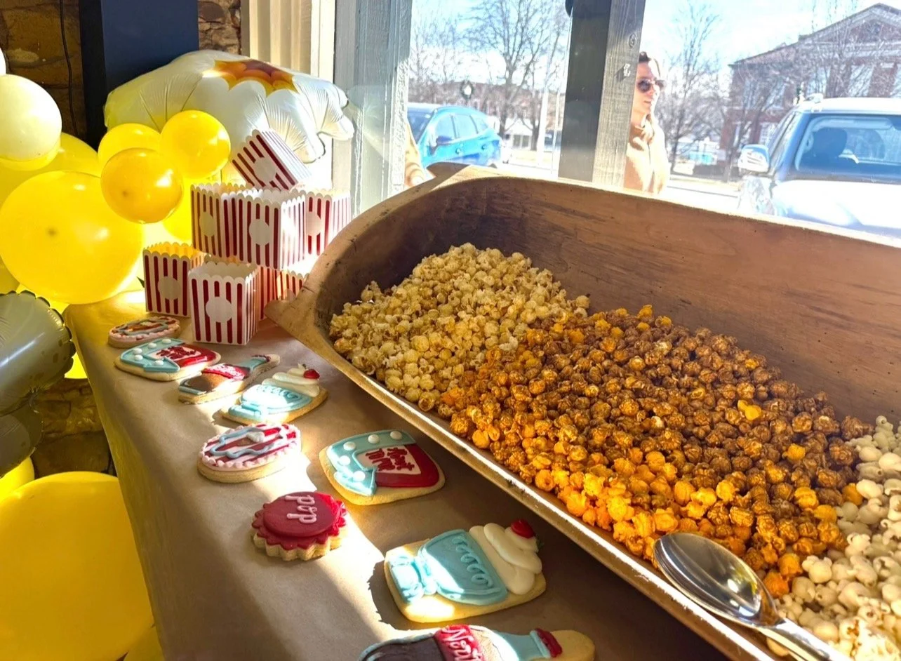 A table with colorful cookies and popcorn, decorated with yellow and white balloons and popcorn-themed containers, near a window showing people outside on a street.