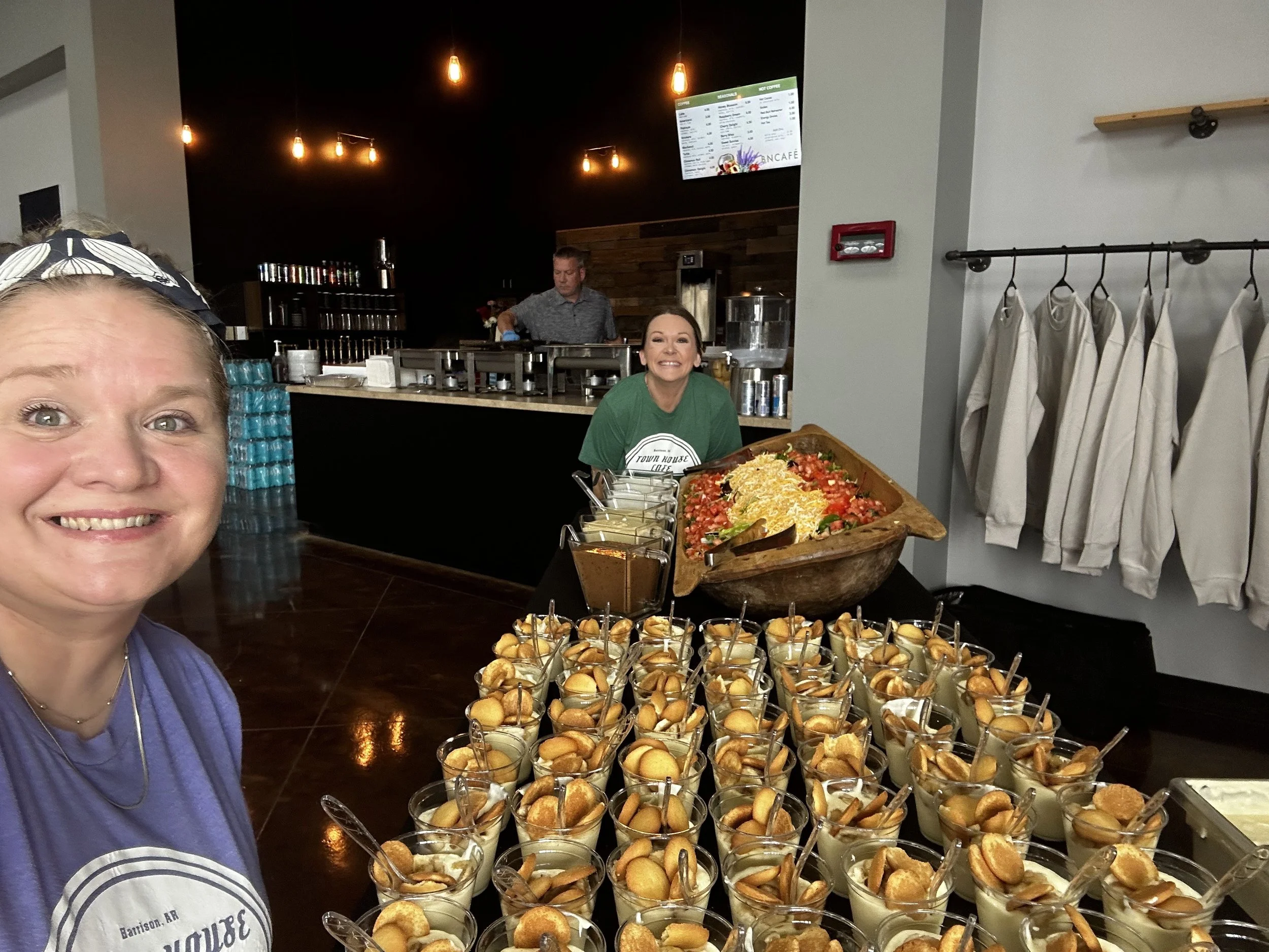 A woman smiling at a food buffet with small cups filled with a dessert topped with cookies, and a large bowl of salad on the table. In the background, another woman stands behind the counter, and a man is working in the kitchen. The decor includes hanging light bulbs and a flat-screen TV on the wall.