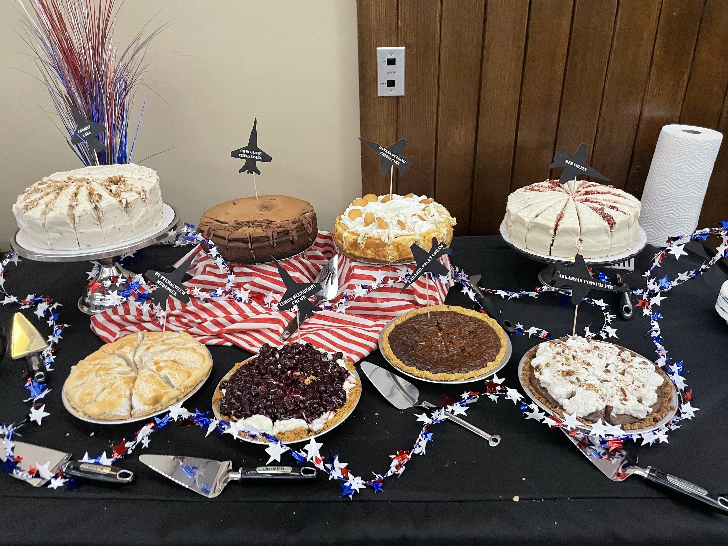 A display of assorted pies on a table decorated with red, white, and blue star-themed decorations, along with a paper towel roll and some pens.