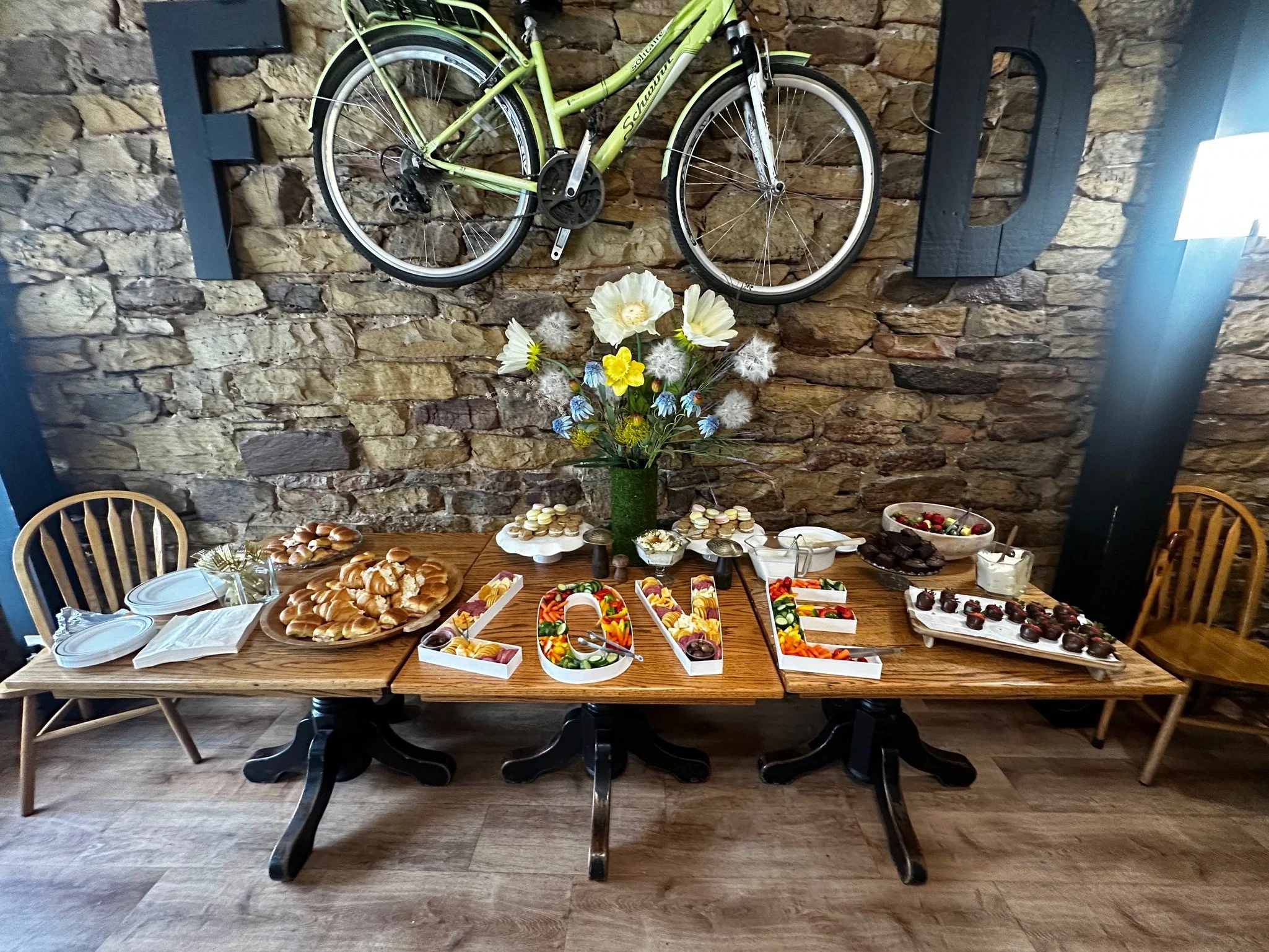 A dining table set for a celebration with various finger foods and desserts, including cookies, cupcakes, fruit, and candy; a large bouquet of flowers in a green vase is at the center of the table. The backdrop features a stone wall with a green bicy