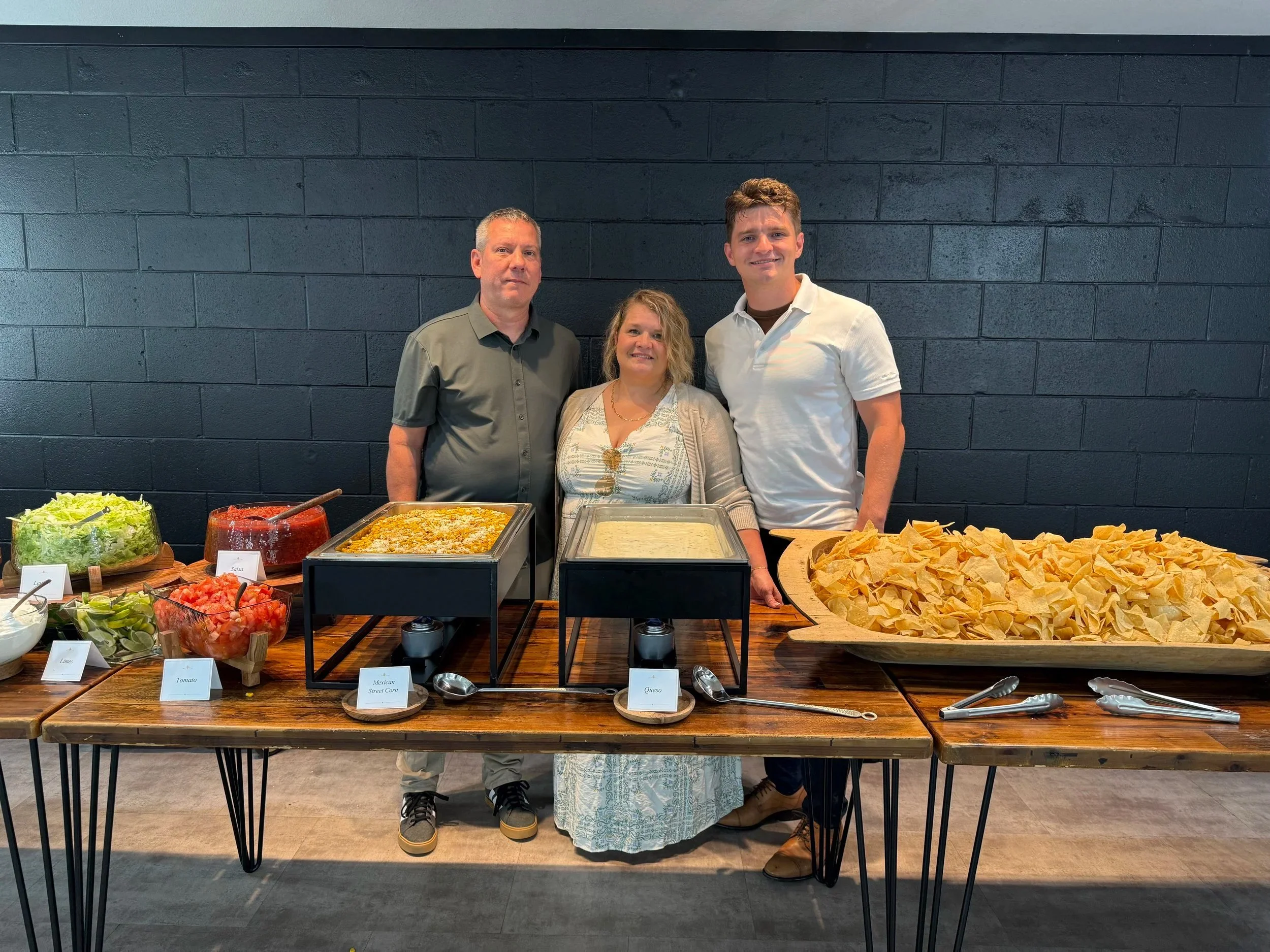Three people are standing behind a buffet table with various food items, including salads, salsa, Mexican street corn, queso, and tortilla chips, set against a dark brick wall.