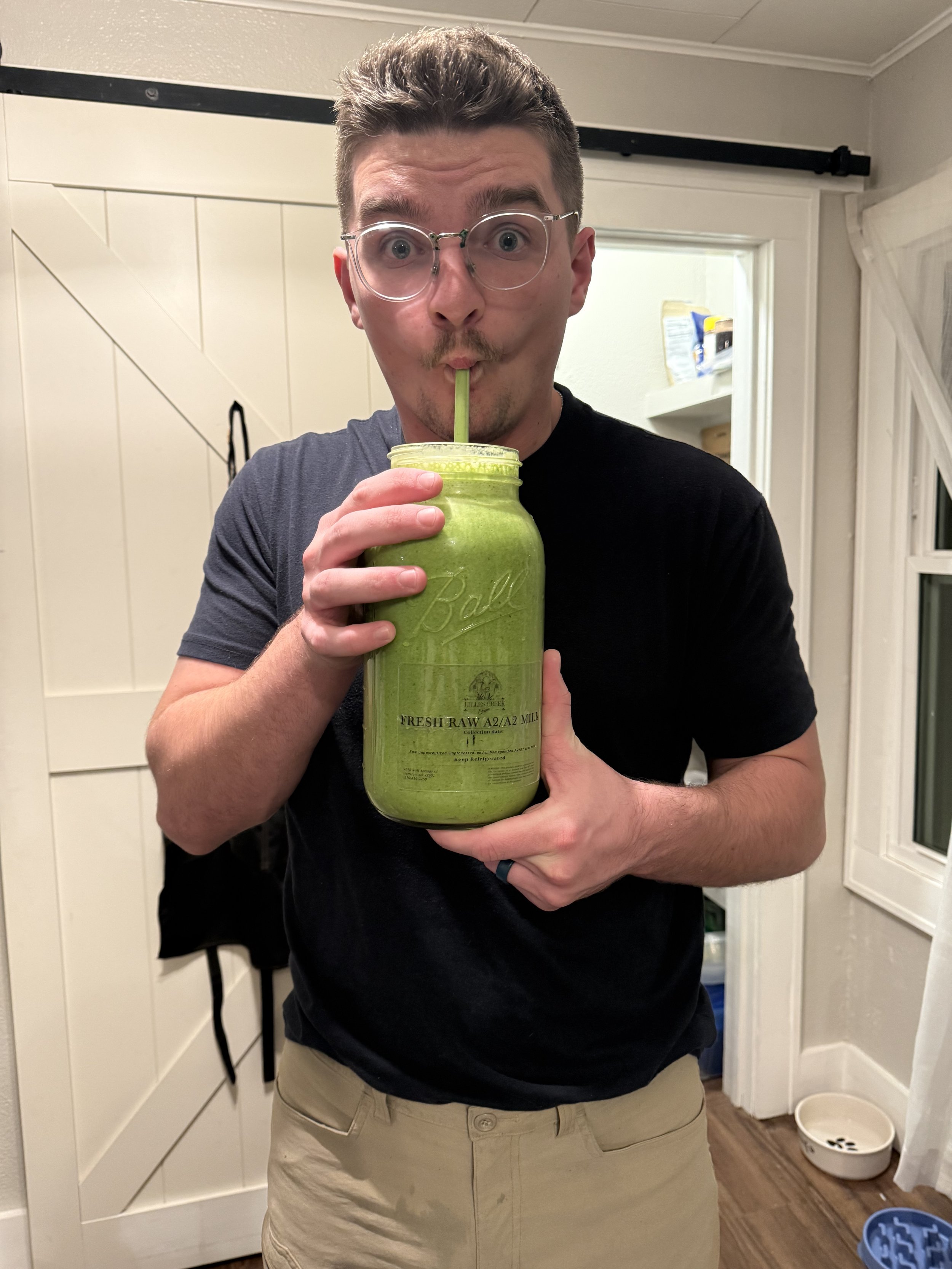 A man wearing glasses and a black t-shirt drinks a green smoothie from a glass jar using a green straw. He is standing indoors in front of a white paneled door.