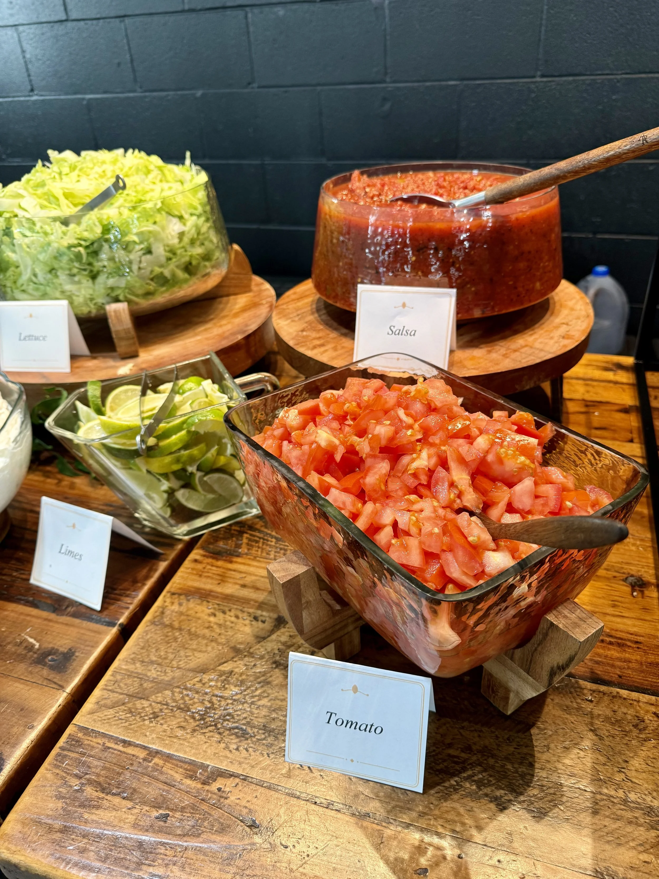A display of various fresh ingredients including chopped tomatoes, sliced limes, shredded lettuce, and salsa in bowls on a rustic wooden table.