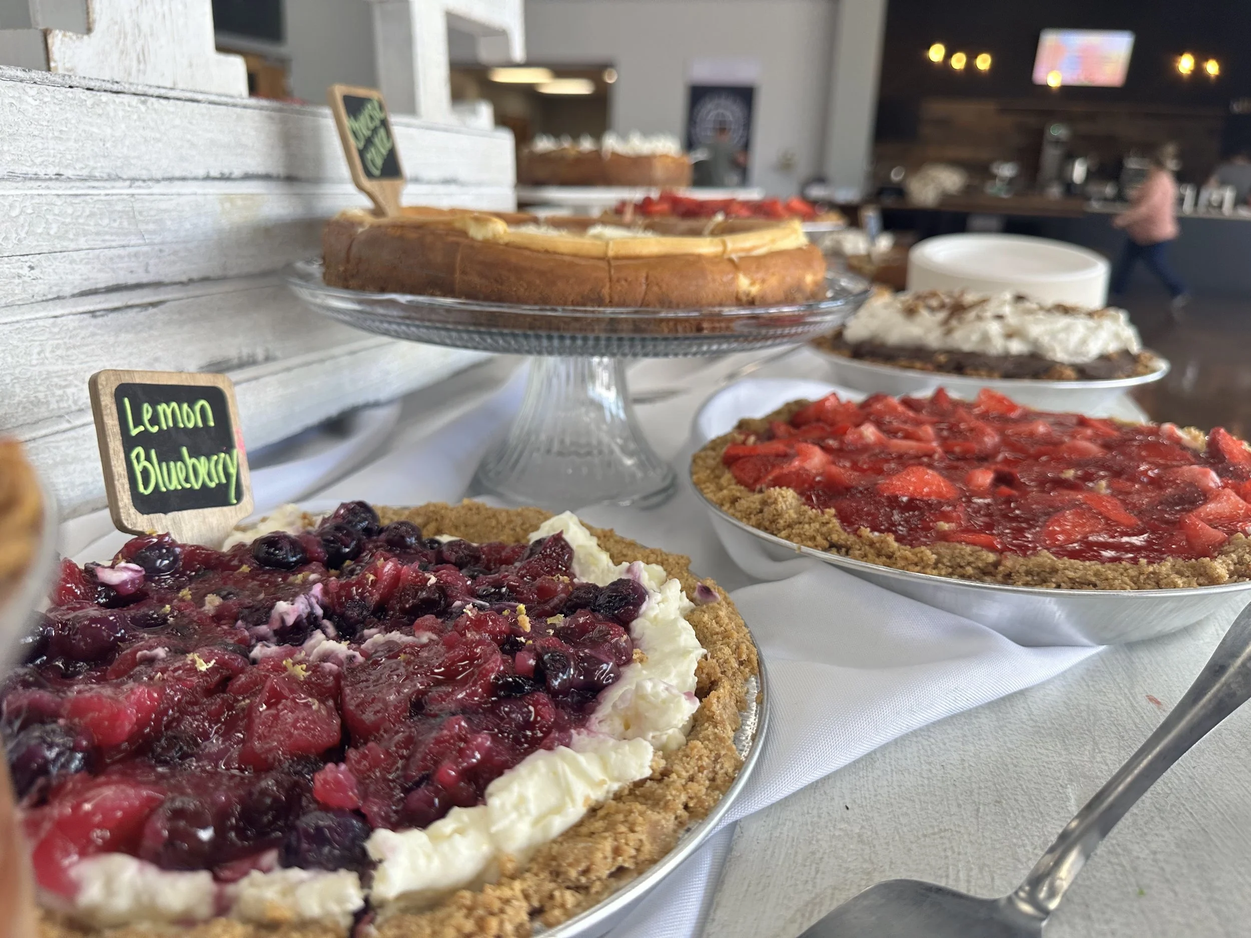 Assorted desserts on a table, including a blueberry crumble pie, a strawberry pie, and a cheesecake with strawberries, displayed on glass and ceramic plates.