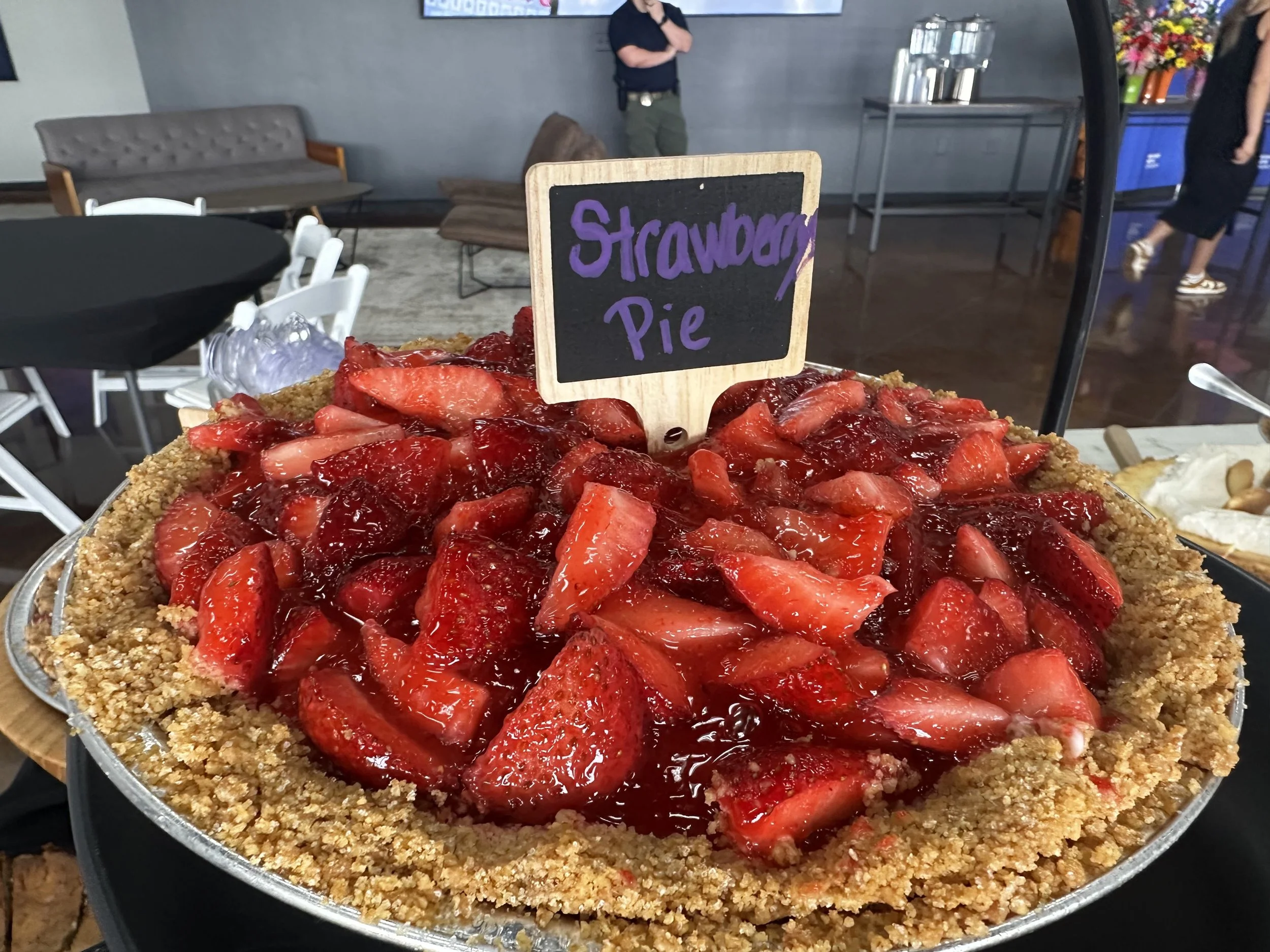 A strawberry pie with a crumb crust and fresh strawberries on top, labeled as 'Strawberry Pie' on a small chalkboard sign.