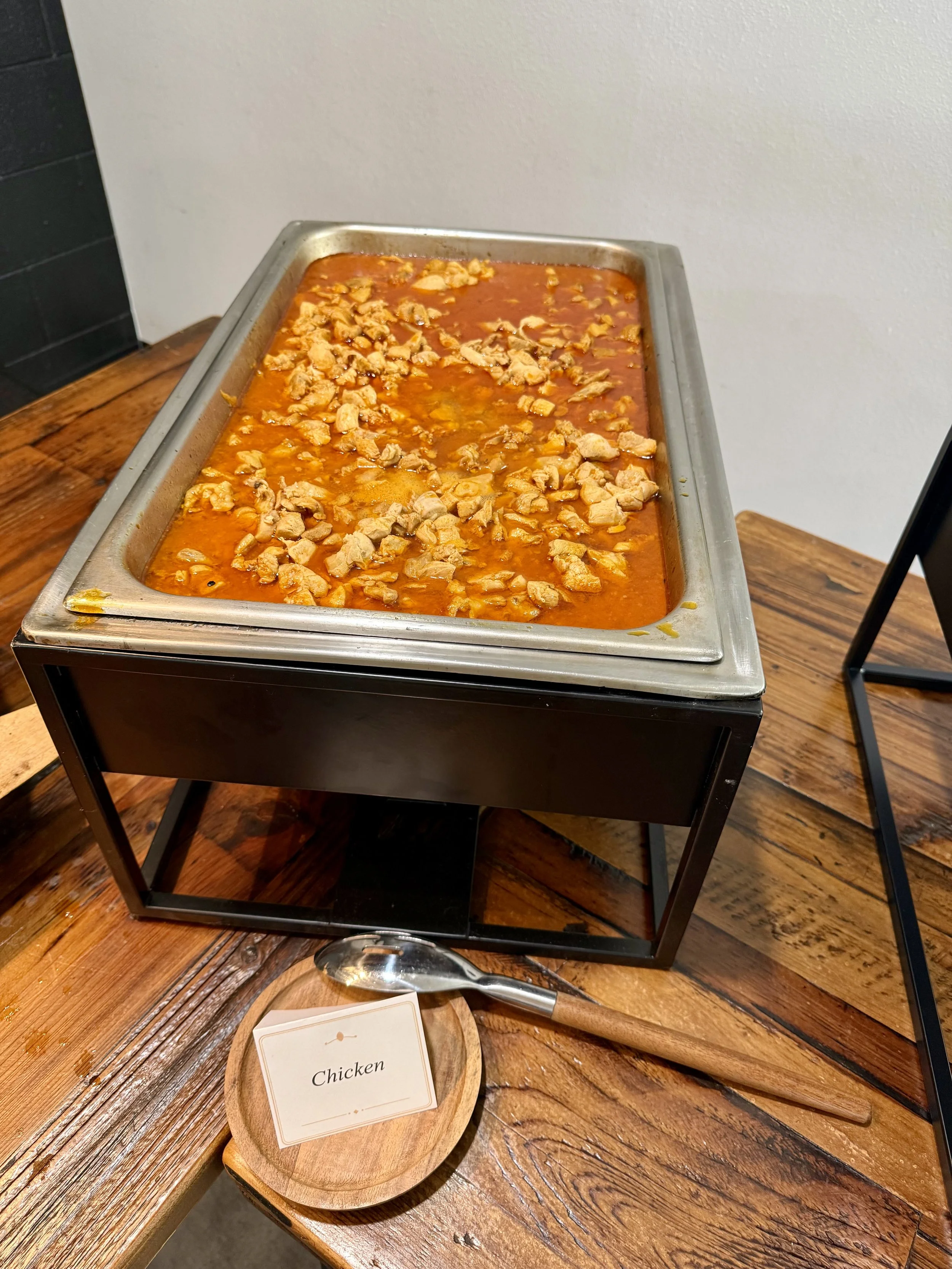 A chafing dish filled with cooked chicken in sauce, placed on a wooden table at a buffet or cafeteria setting. A small wooden plate with a label that reads 'Chicken' and a serving spoon are in front of the dish.