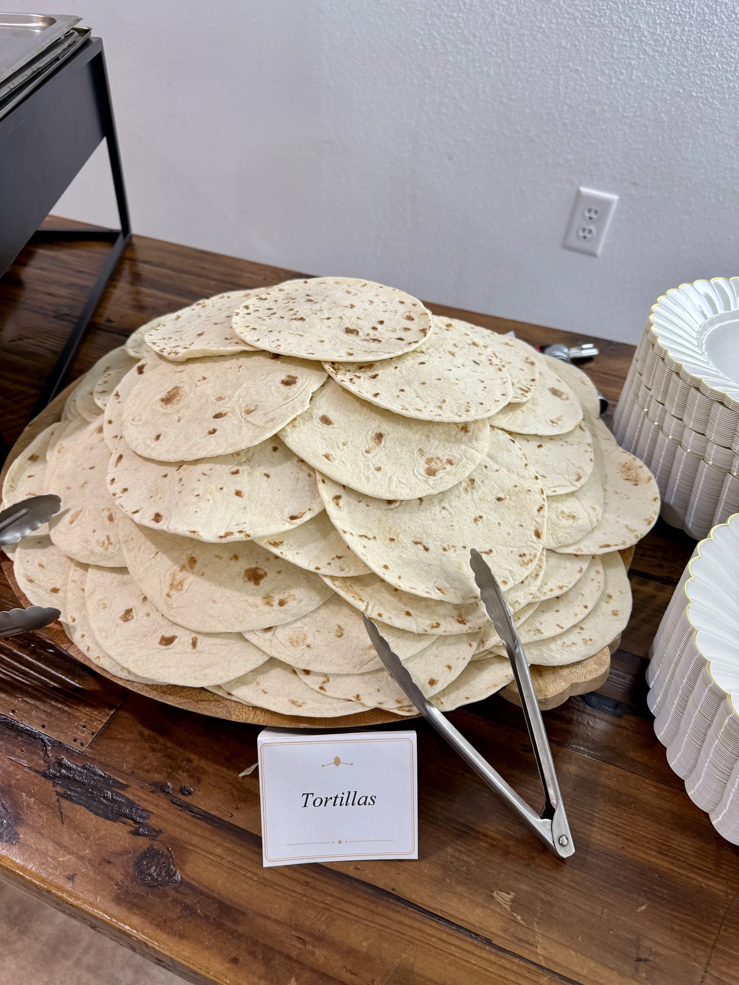 A large pile of tortillas on a wooden table, with serving tongs and a sign labeled 'Tortillas'.