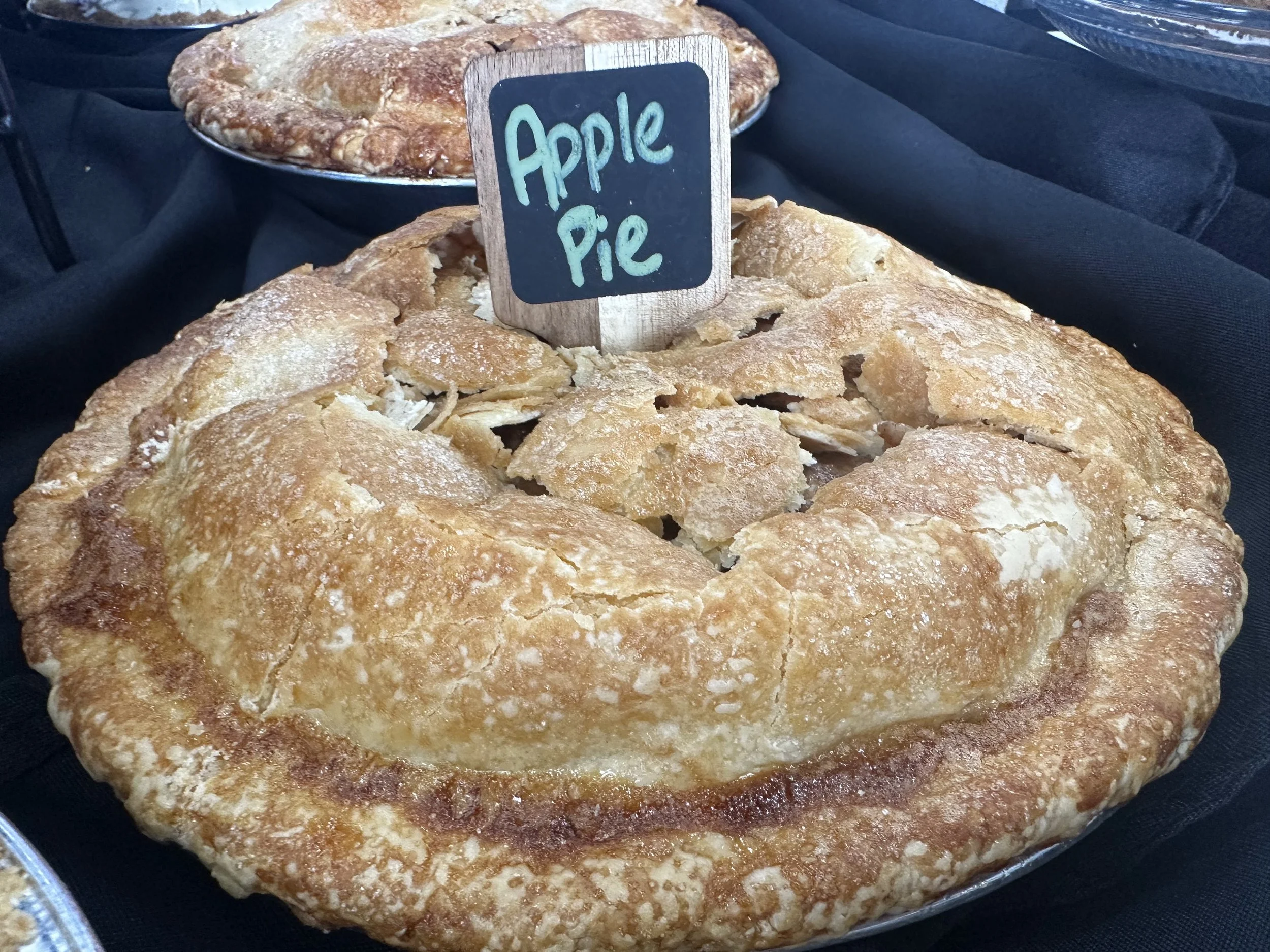 Close-up of a freshly baked apple pie with golden crust and a small chalkboard sign reading 'Apple Pie' placed on top. The pie is displayed on a black surface with other baked goods in the background.