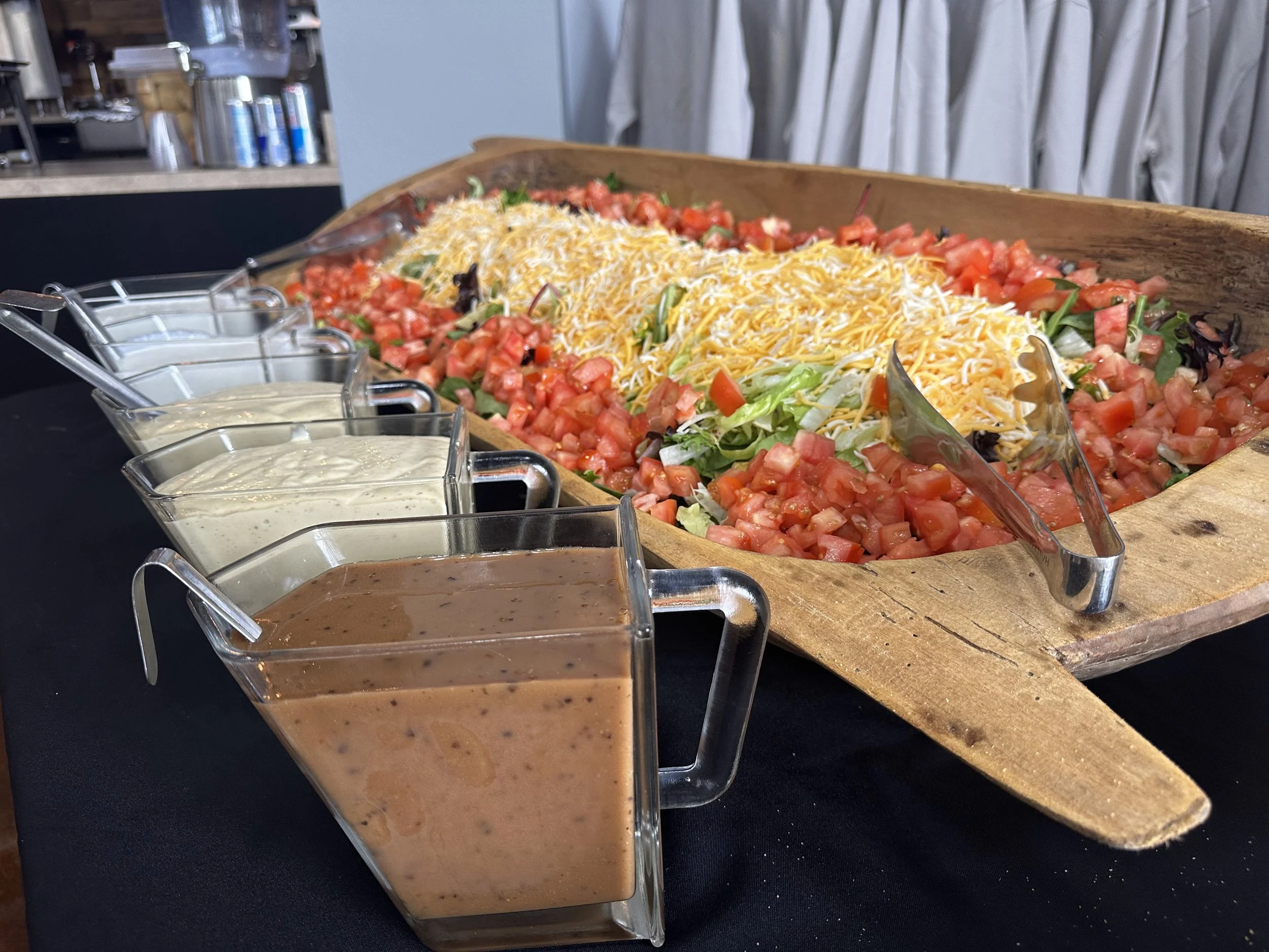 A wooden tray filled with chopped tomatoes, shredded cheese, and leafy greens. In front of the tray, there are four small glass containers with different salad dressings.