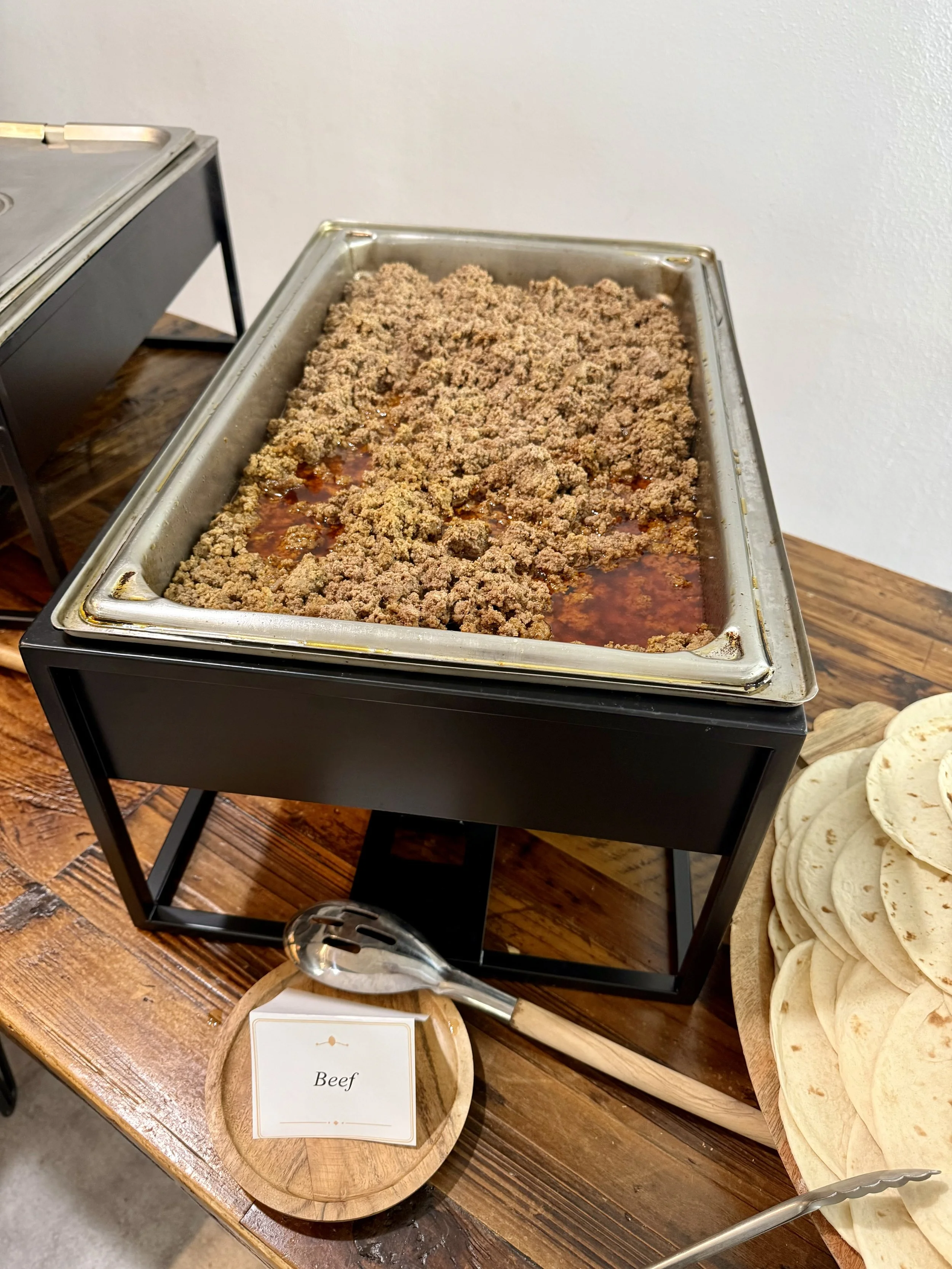 Tray of cooked ground beef with a small sign in front labeled 'Beef', set on a wooden table with a plate of tortillas nearby.