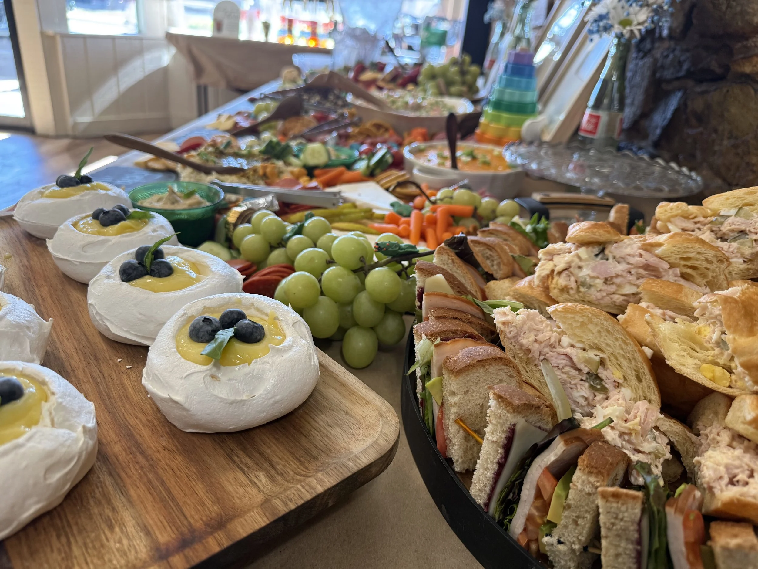 A buffet table with various appetizers including small pavlova desserts topped with blueberries, fresh green grapes, a vegetable platter with carrots, celery, and tomatoes, bowls of dips, and an assortment of sandwiches and rolls, set in a bright, co