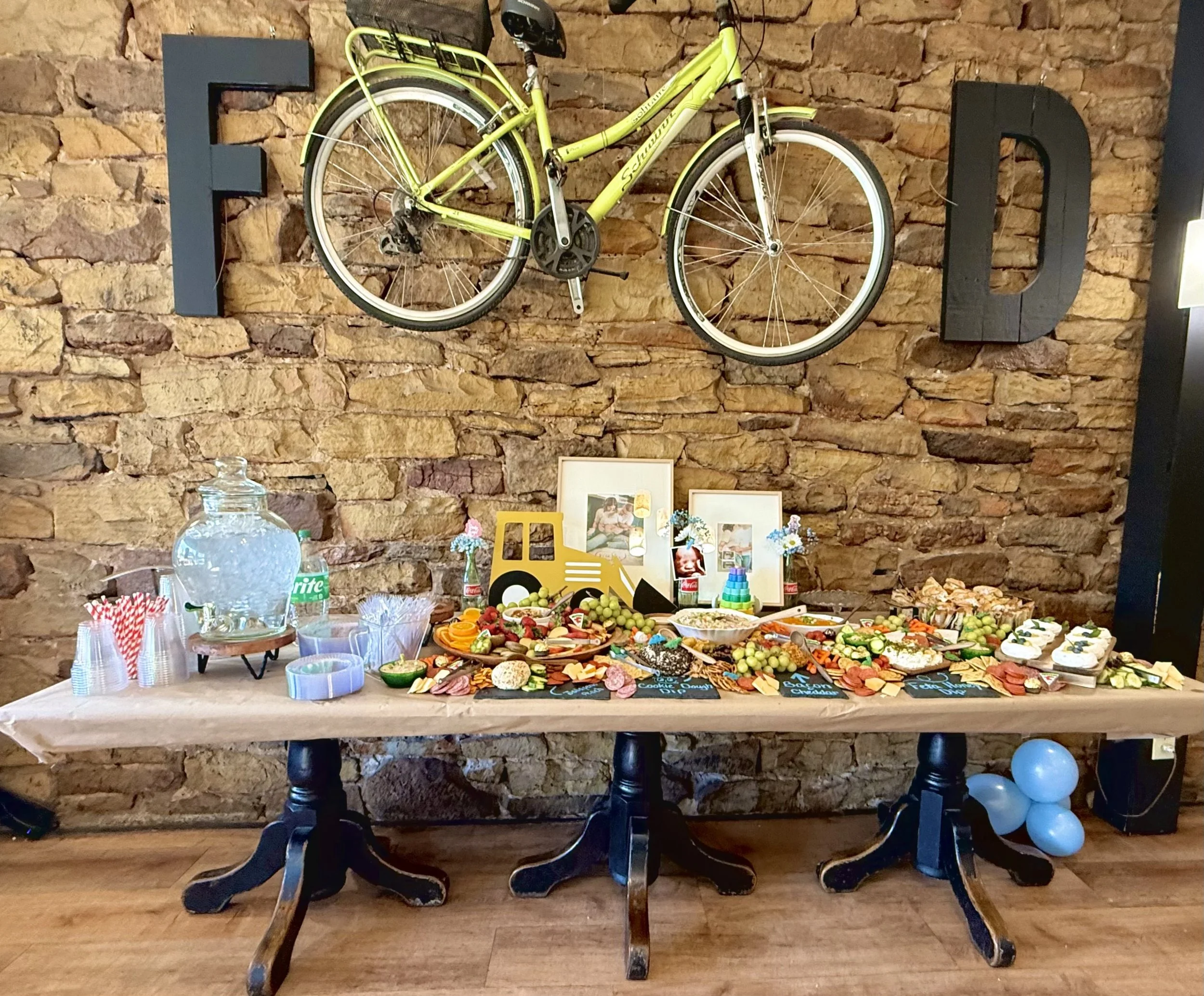 A buffet table set for a celebration with a variety of food and snacks, featuring a brick wall background with large decorative letters 'F' and 'D', a yellow bicycle mounted on the wall, and framed photographs.
