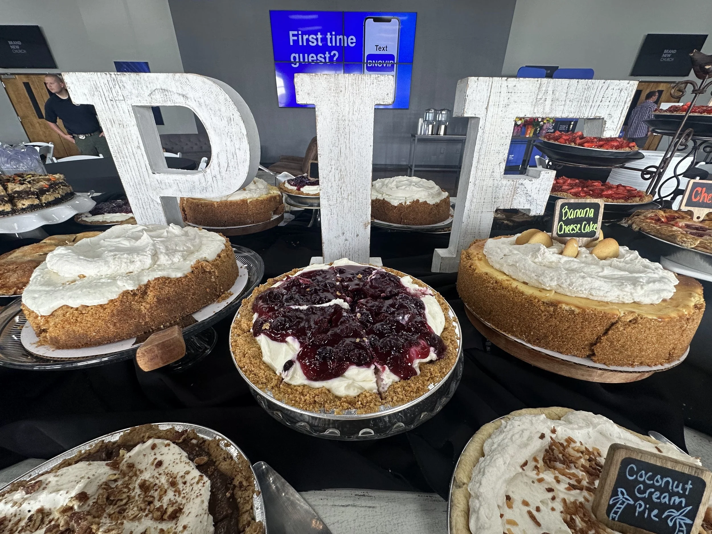 A table of various pies with different flavors, including banana cheesecake and coconut cream, at a bakery or tasting event. Large white decorative letters spell out 'DTE' in the foreground, with a screen in the background displaying a sign about fir