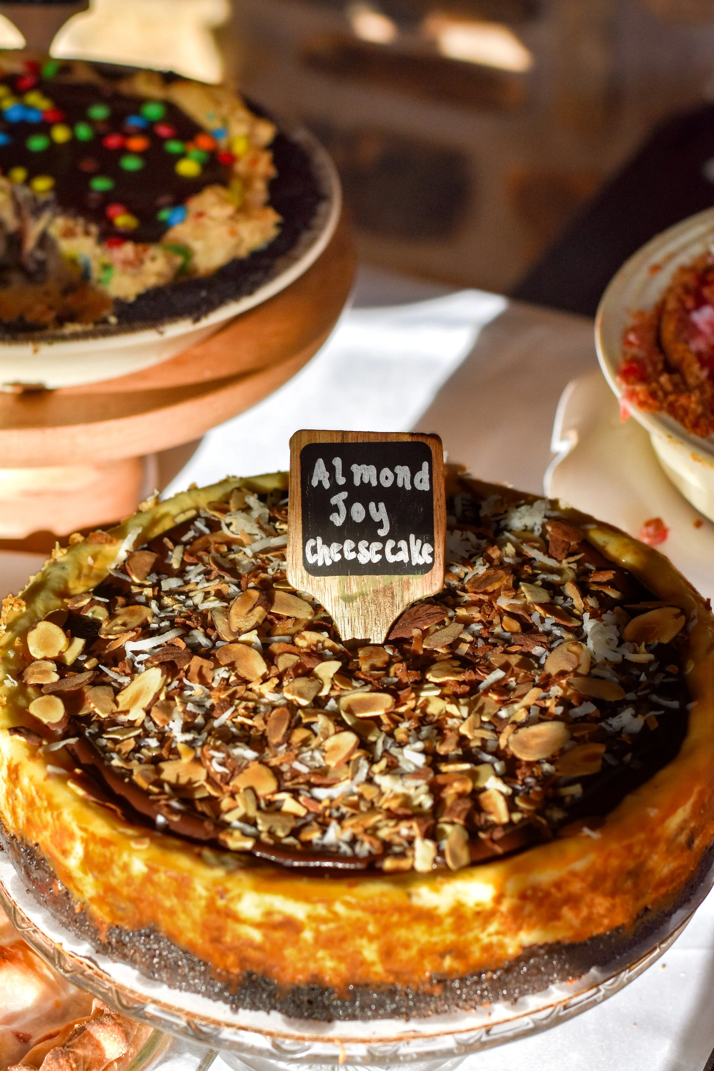 A close-up of a cheesecake topped with sliced almonds and dark chocolate, with a small chalkboard sign reading 'Almond Joy Cheesecake.'