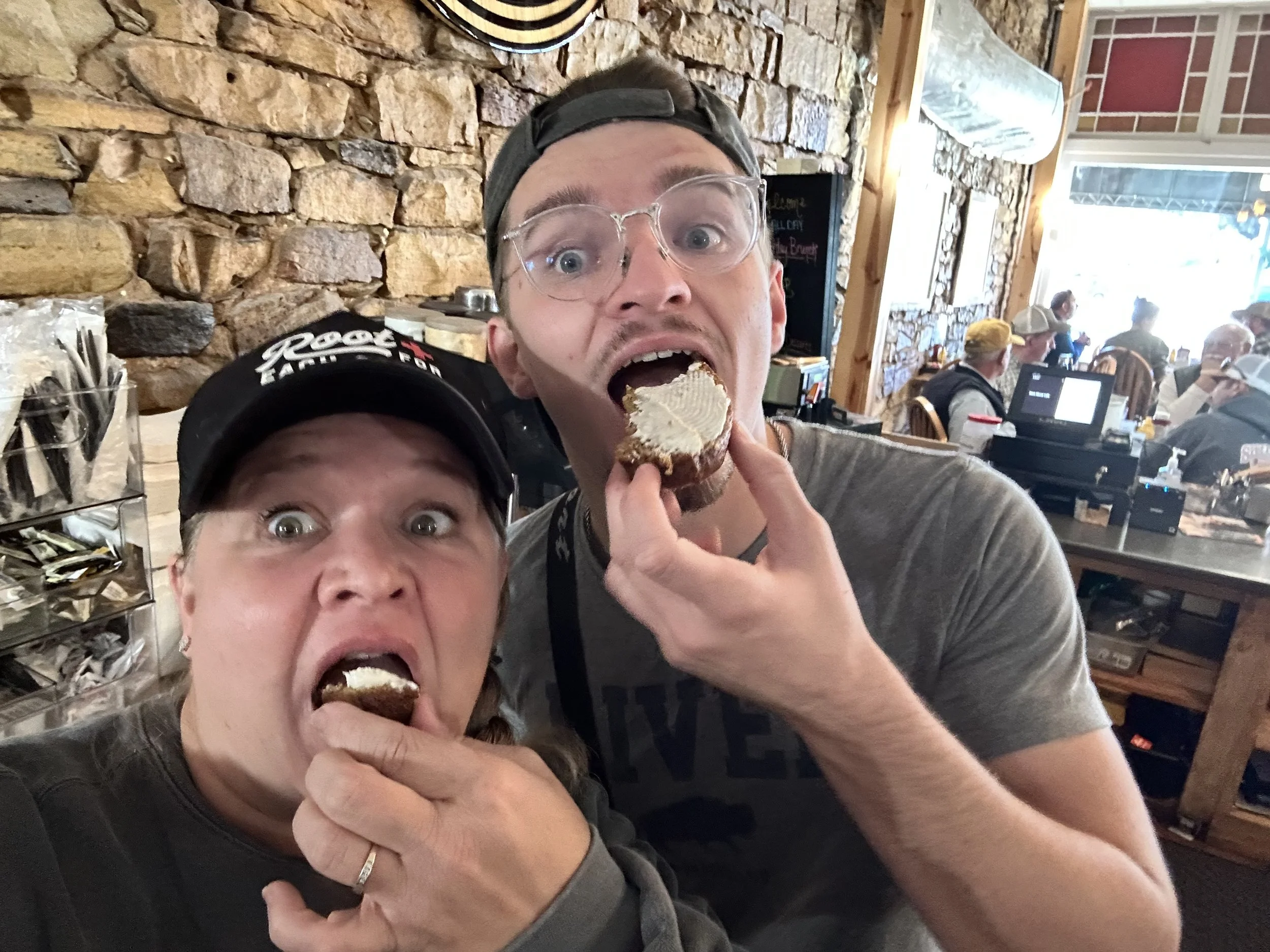 A woman and a man are taking a selfie inside a restaurant. Both are covering their mouths with their ice cream cones, with their eyes wide open, expressing excitement or surprise. The restaurant has a rustic decor with a stone wall and several other patrons in the background.