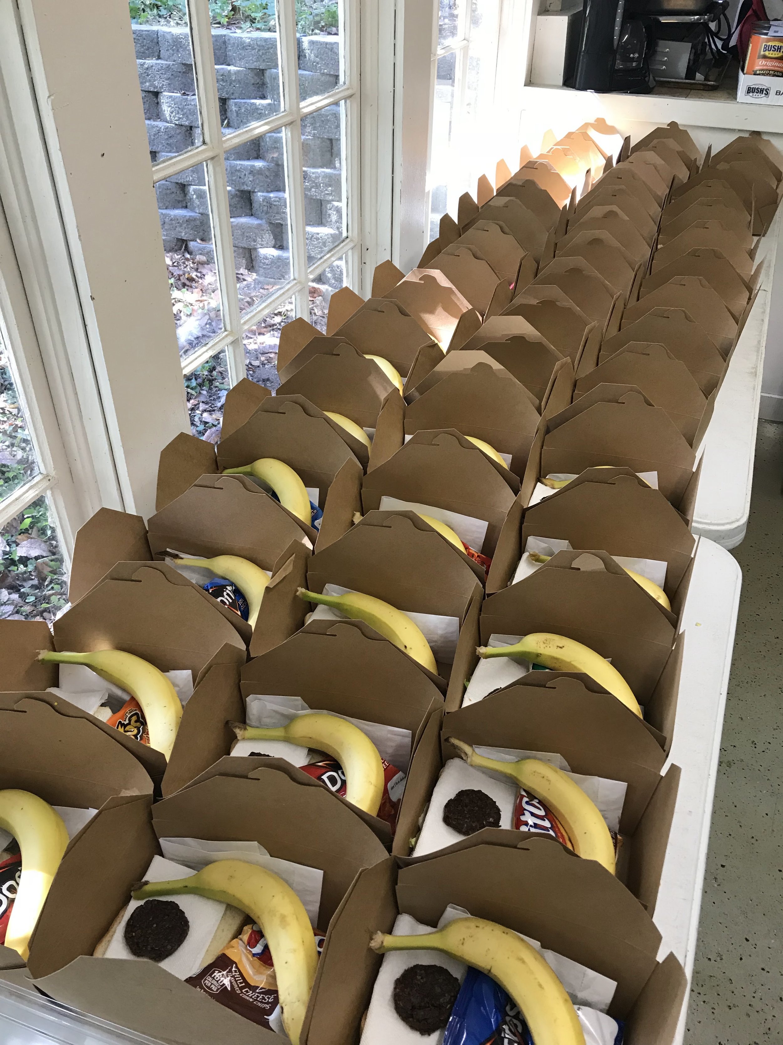 Multiple brown paper grocery bags filled with bananas, Oreos cookies, and other snacks lined up on a white table near a window.