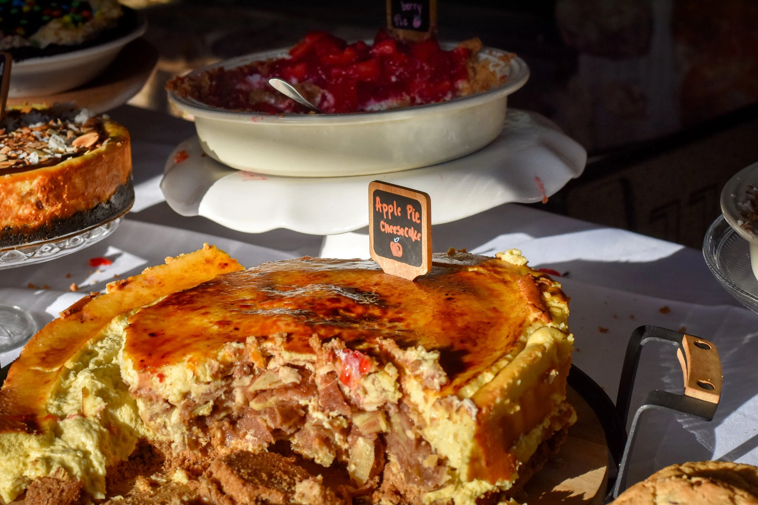 A slice of apple pie cheesecake with a crumbly crust, topped with a gold-brown layer, on a table at a bakery display.