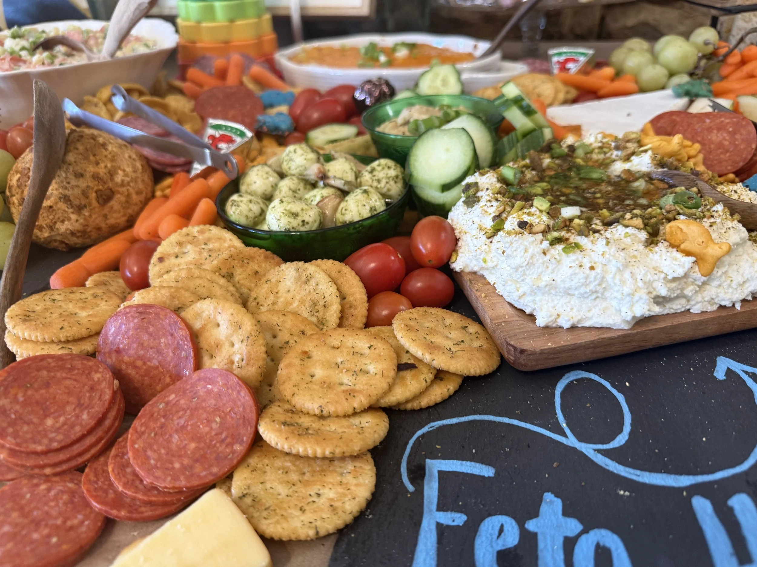 An assortment of snack foods including pepperoni slices, crackers, cherry tomatoes, cucumber slices, cheese, and bowls of dip on a wooden serving board.