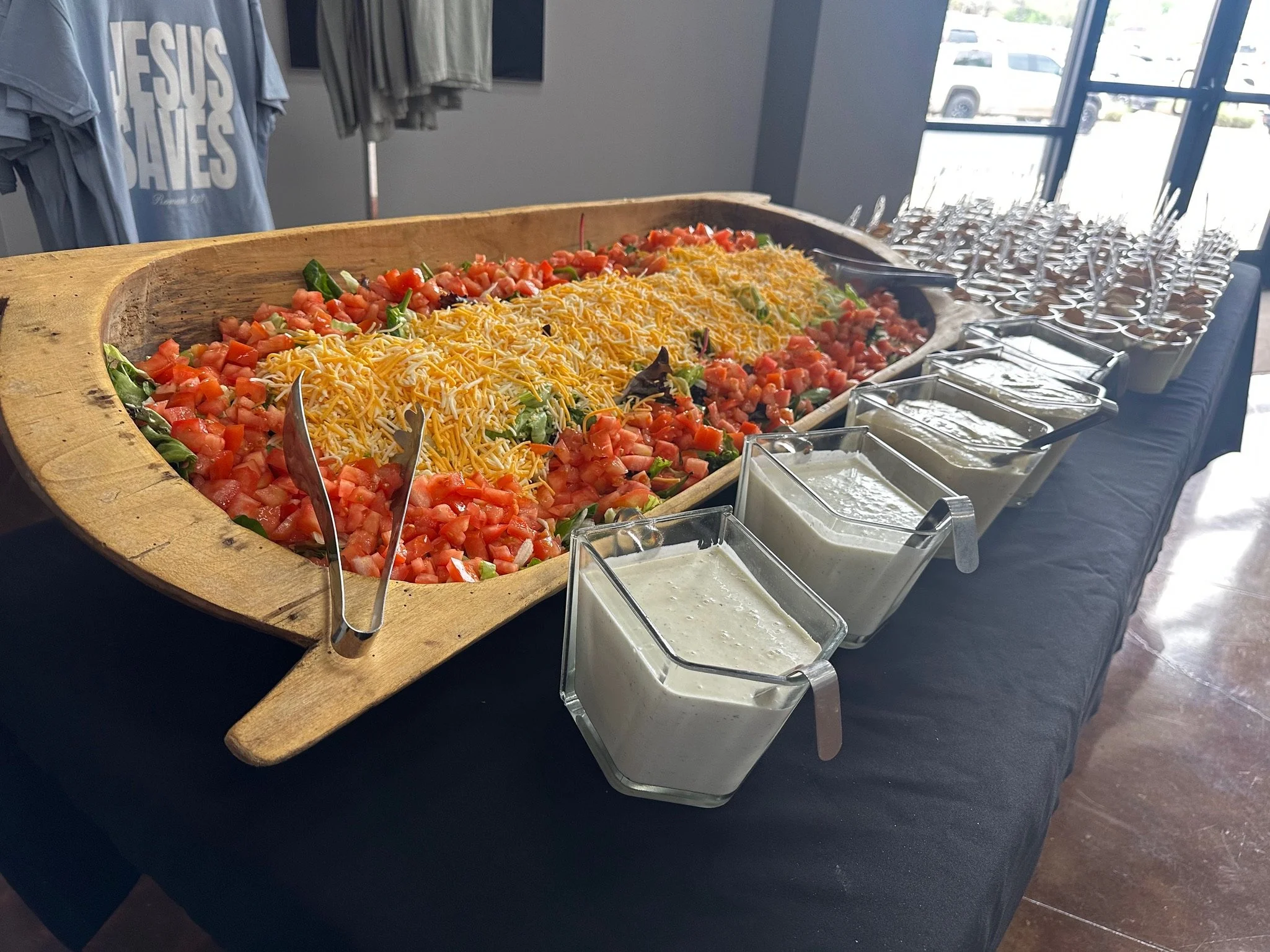 A table with a large wooden bowl of chopped tomatoes, shredded cheese, and salad greens, along with smaller containers of dressing and toppings, set up for a salad bar in a bright room with large windows.