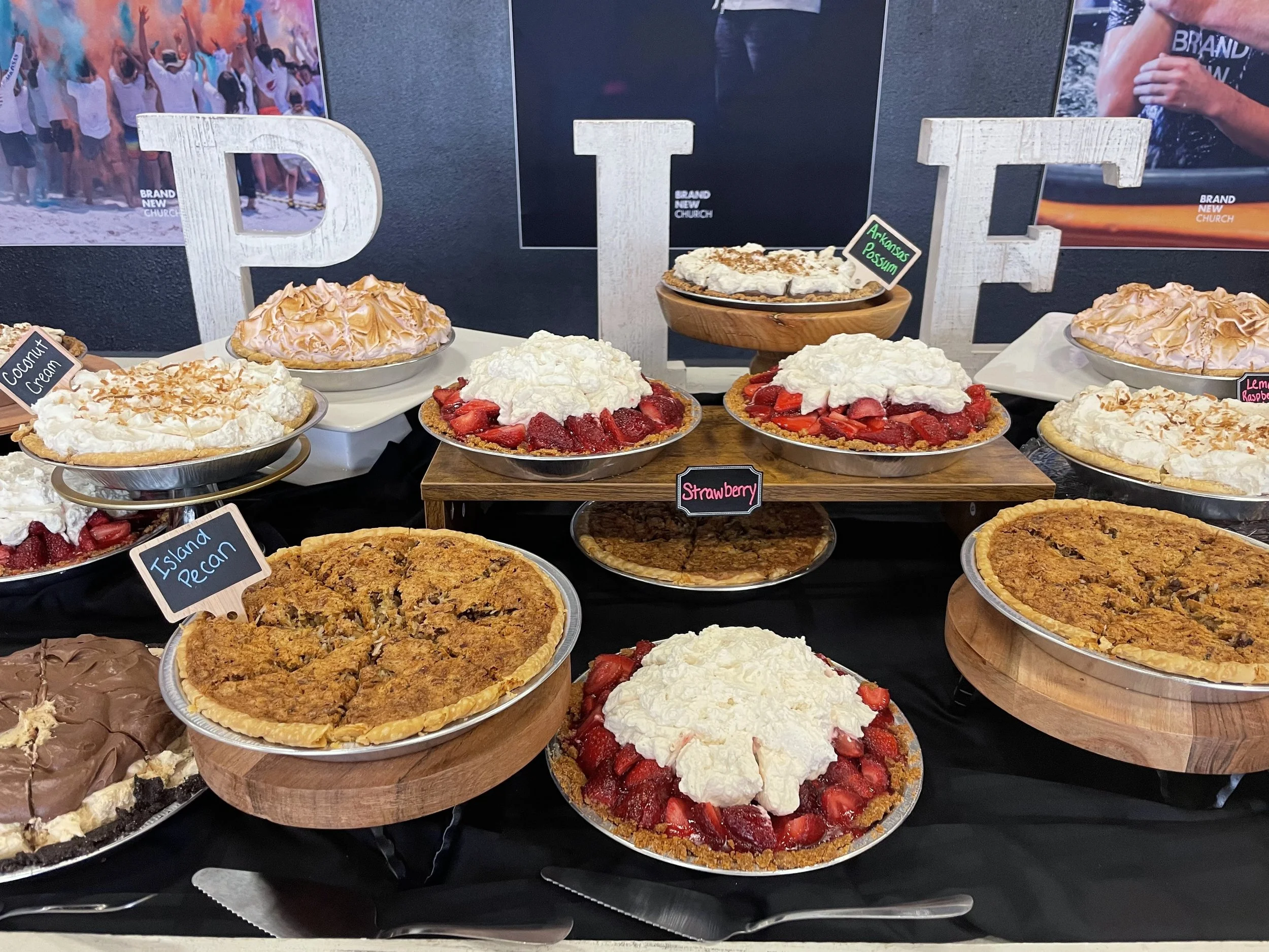 A variety of desserts including strawberry, coconut cream, and lemon raspberry pies displayed on a table, with decorative letters spelling 'P I E' behind them, and posters of people in the background.