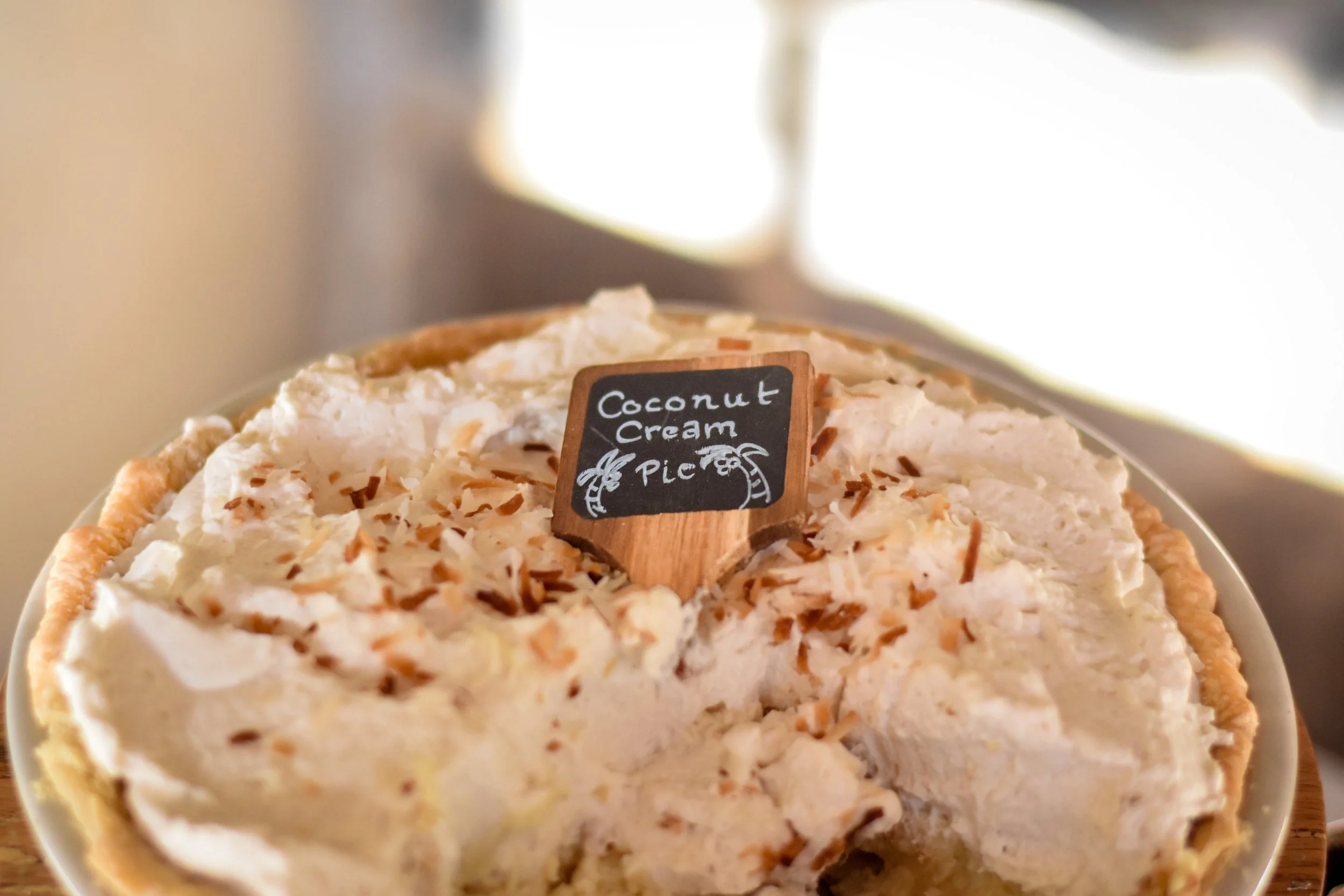 A pie with coconut cream filling topped with toasted coconut flakes and a small chalkboard sign reading 'Coconut Cream Pie' with a pineapple illustration.