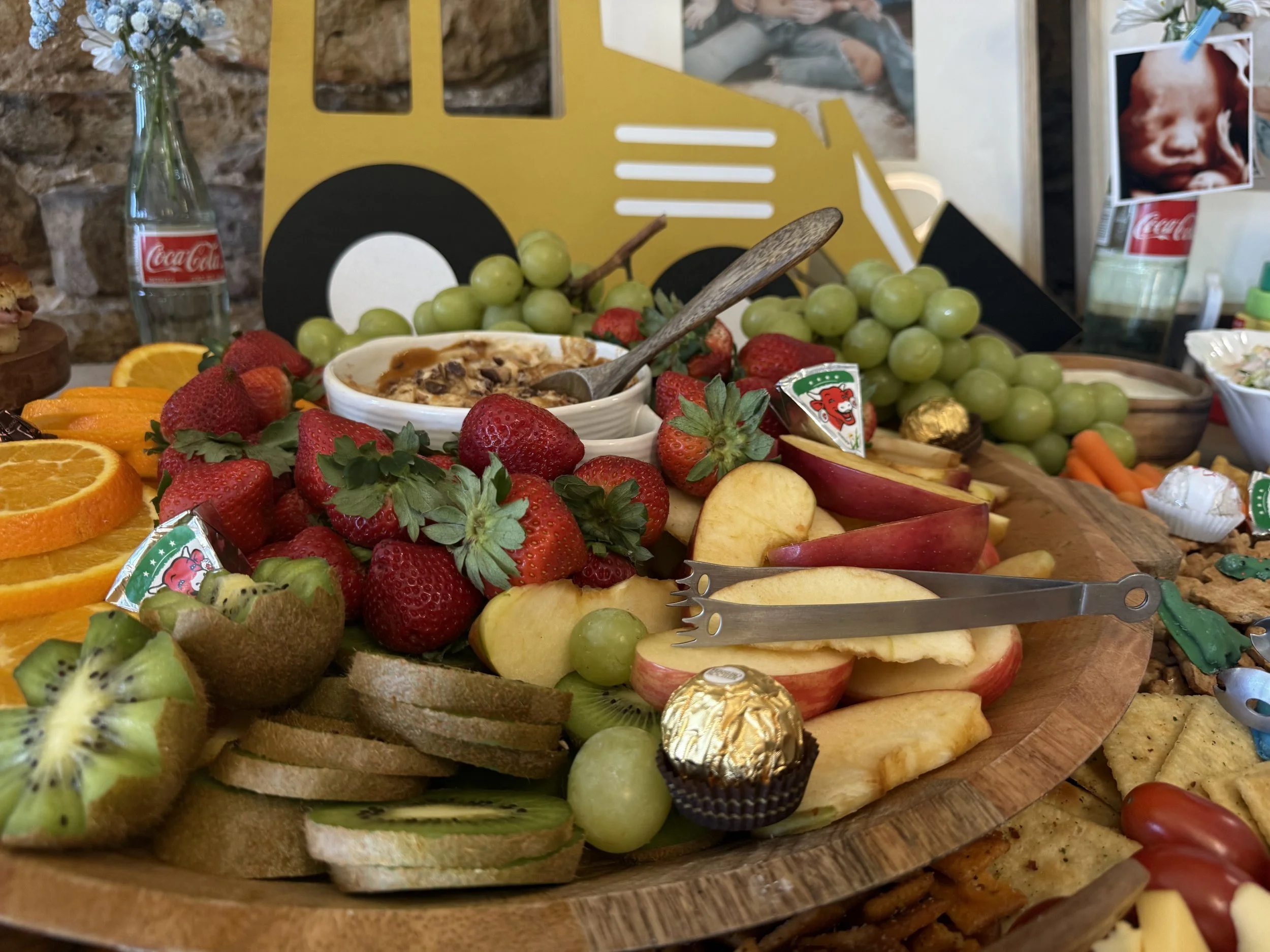 A wooden platter holding grapes, strawberries, oranges, apple slices, kiwi, and various snacks, on a rustic background with a yellow decorative truck.