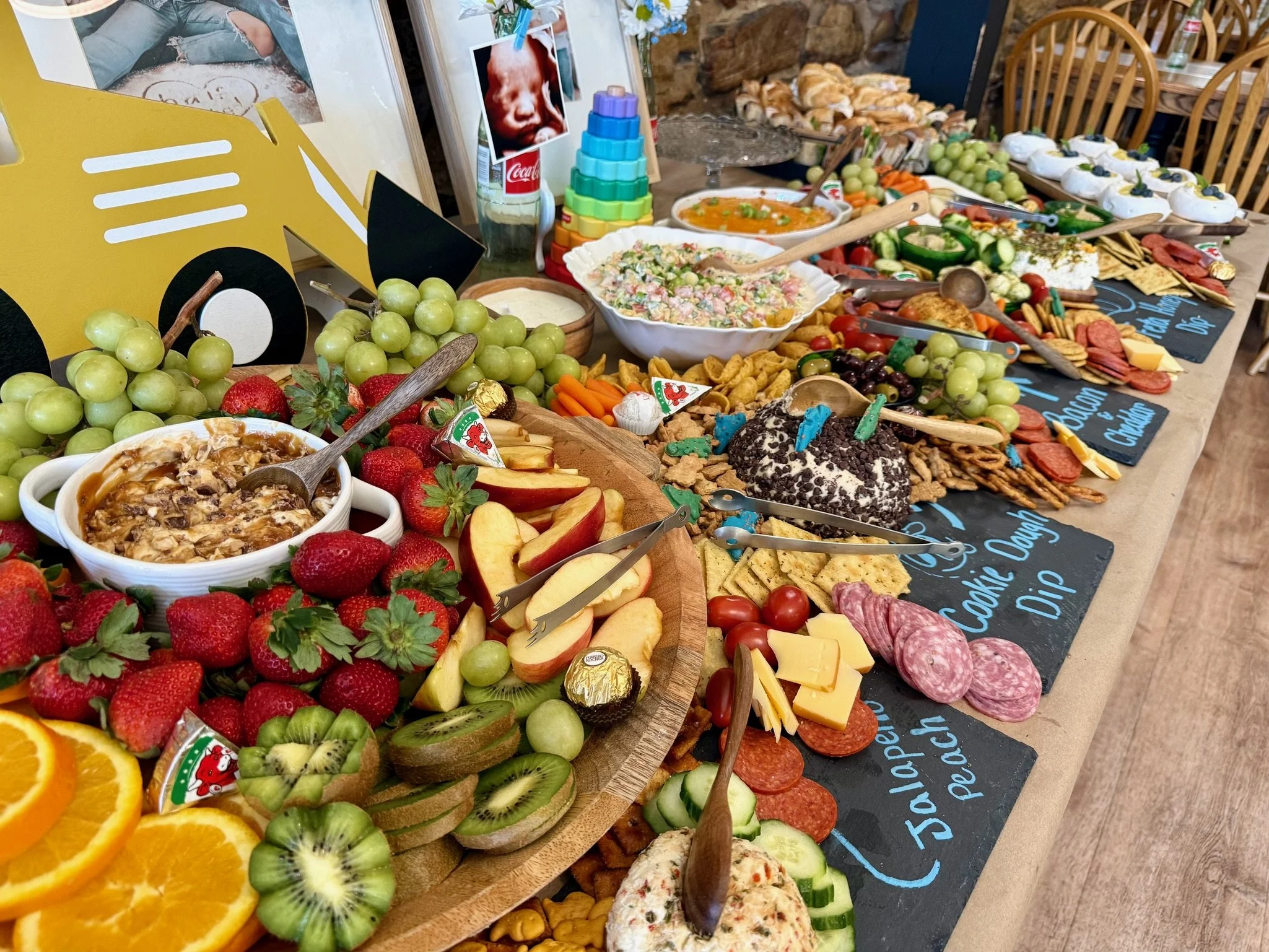 A colorful spread of food and snacks on a table, including grapes, strawberries, oranges, kiwi, sliced apples, cheese, crackers, deli meats, salad, dips, and desserts, with a backdrop of chairs and a stone wall.