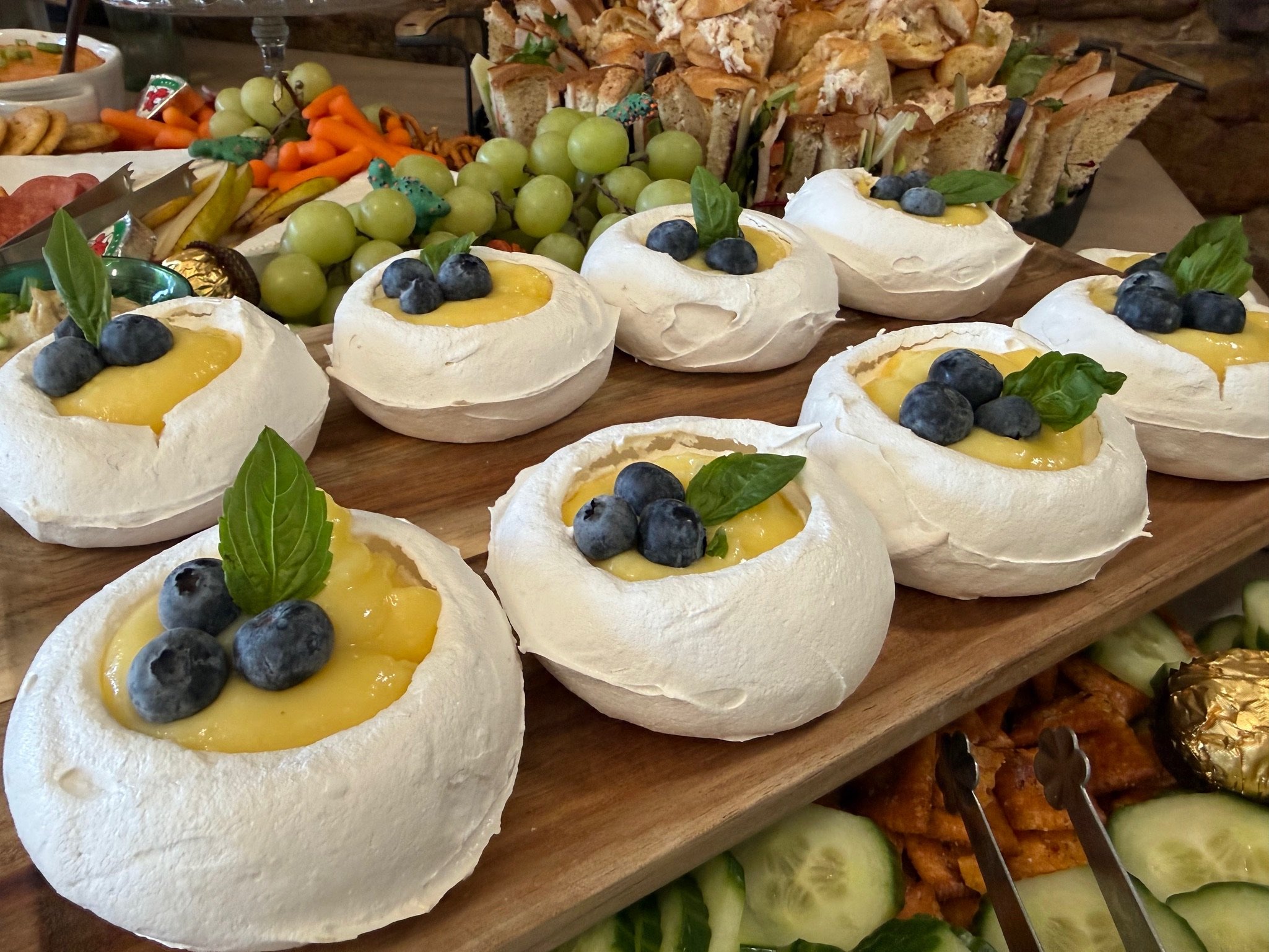 A platter of baked Brie cheese topped with blueberries and basil leaves, with a background of grapes, bread, and other snacks on a wooden table at a buffet.