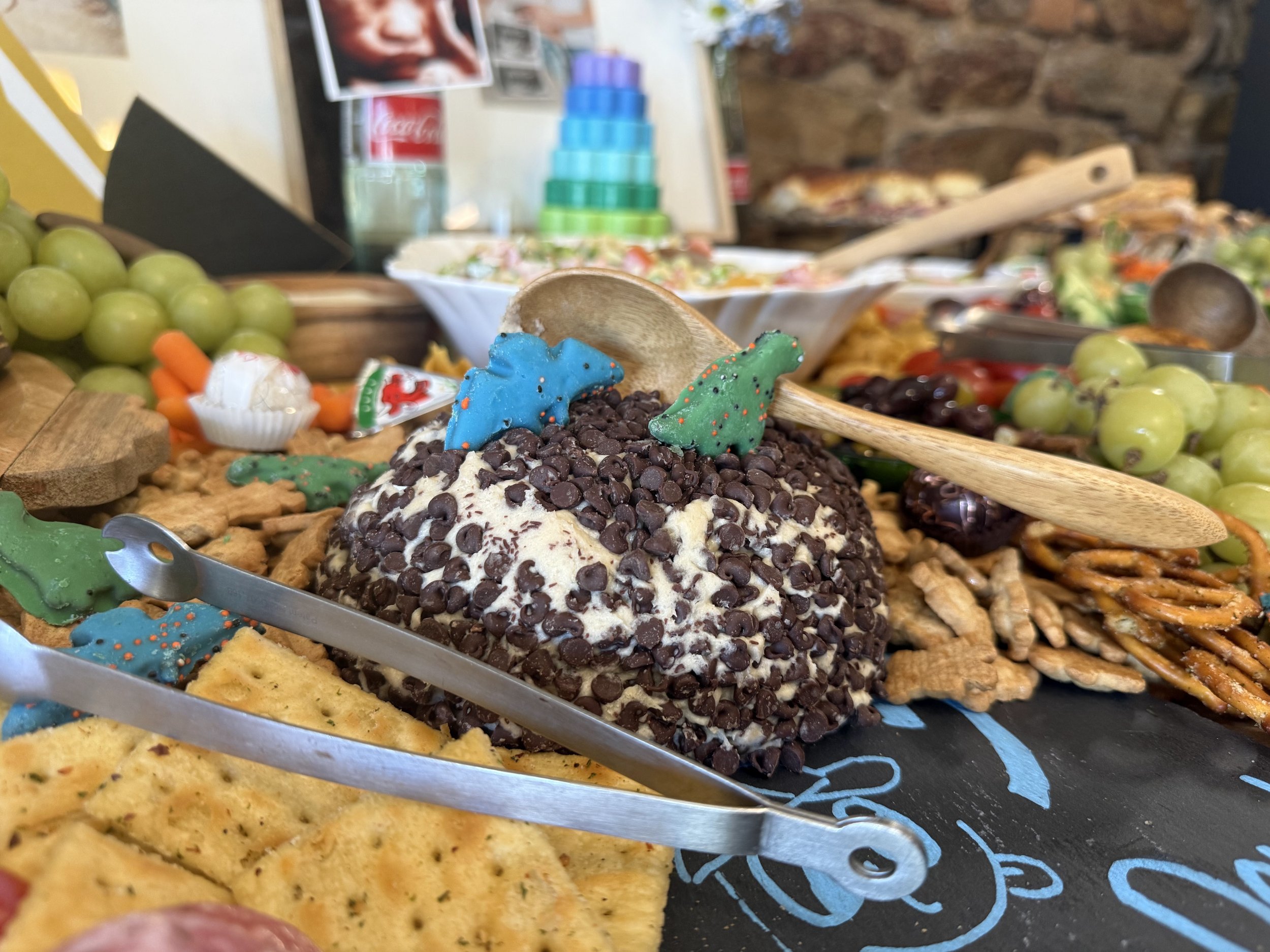 Close-up of a dessert topped with mini chocolate chips and colorful holiday-themed decorations, with other food items and a colorful stacking ring toy in the background.