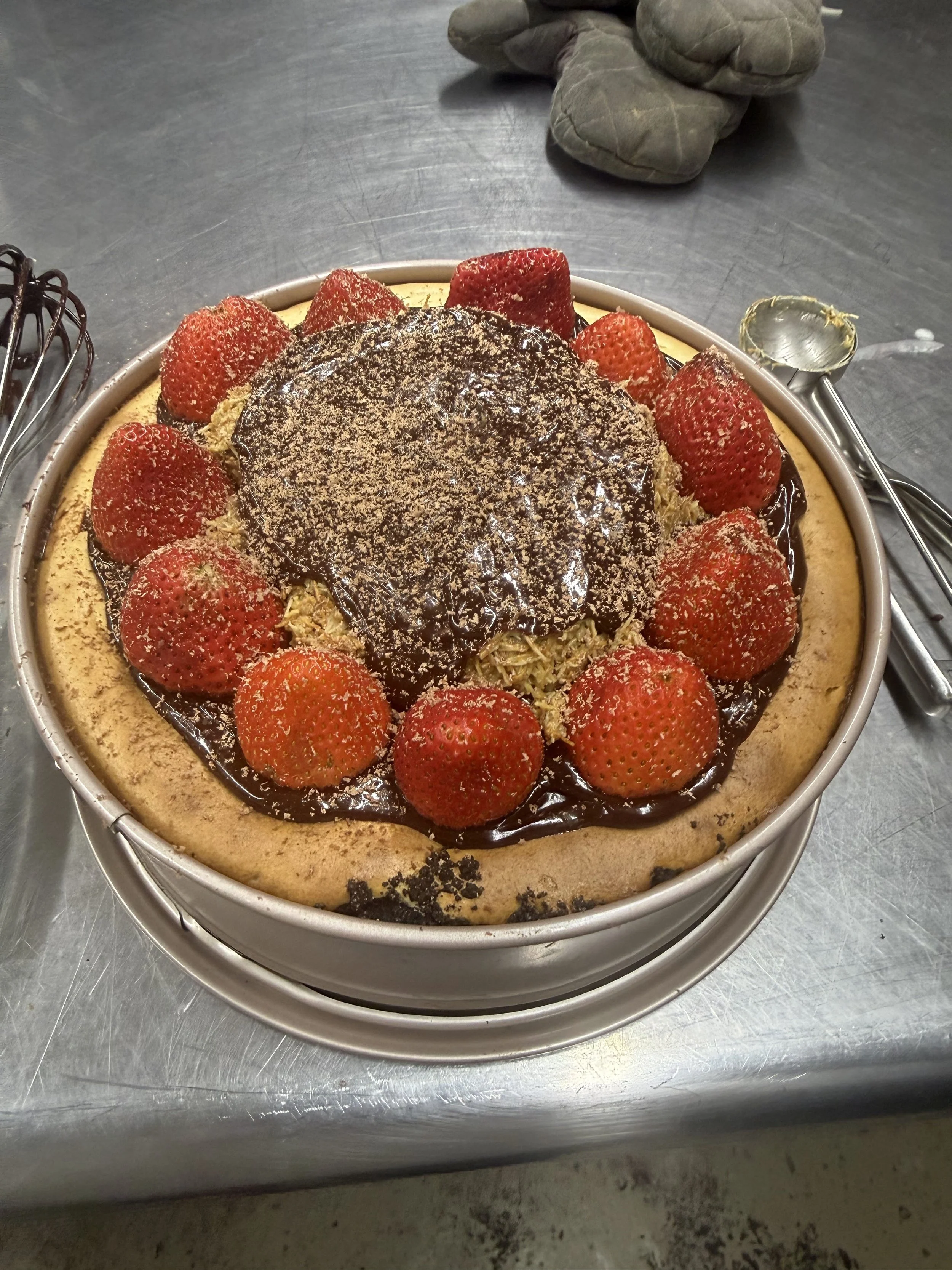 A chocolate dubai pecan cheese cake topped with chocolate glaze, strawberries, and shredded toppings in a round pan on a metal table.