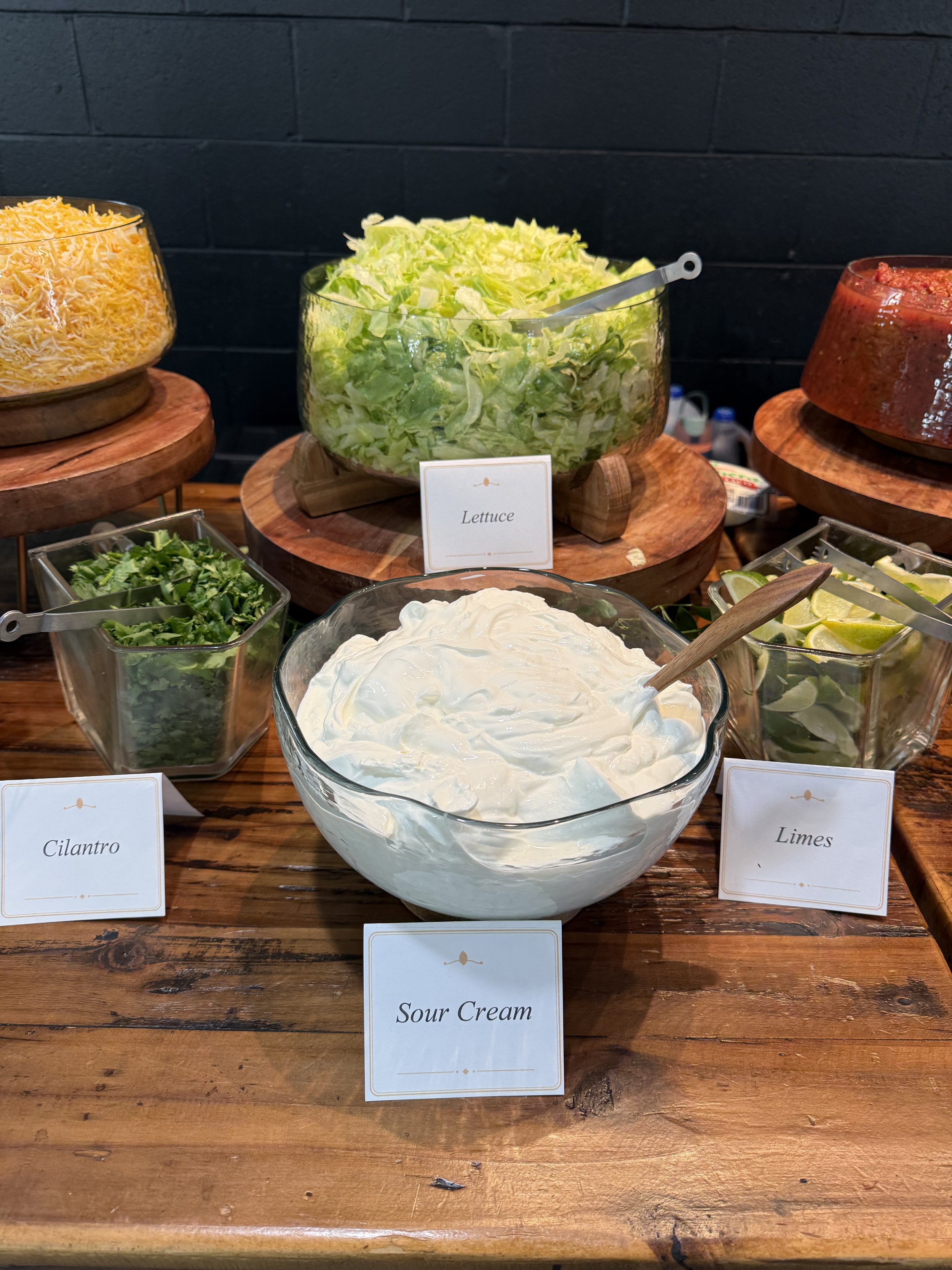 A buffet table with bowls of shredded cheese, shredded lettuce, chopped cilantro, sour cream, and lime wedges, each labeled with small signs.