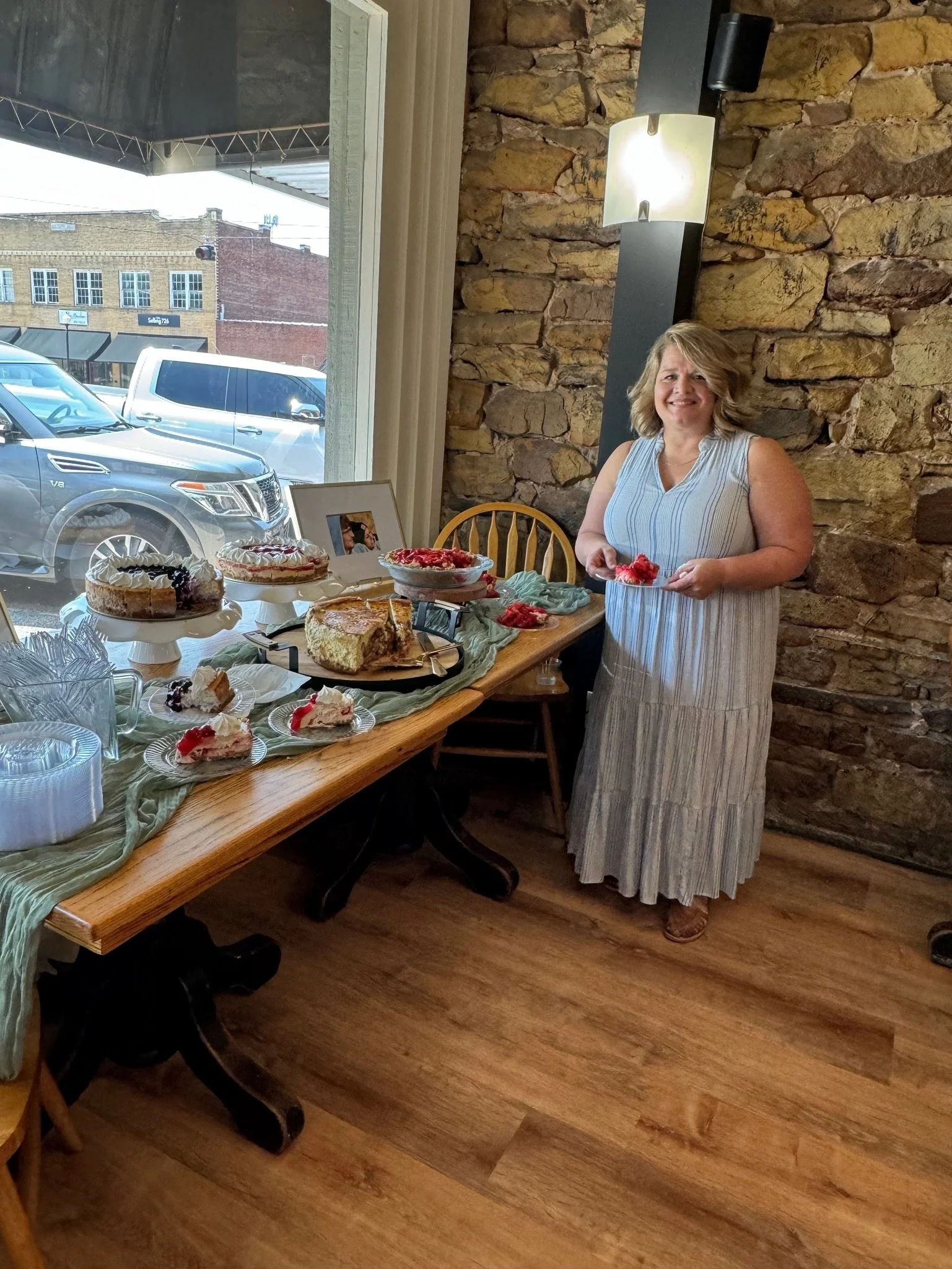 A woman standing by a table with assorted desserts, including pies and cake slices, inside a rustic room with a brick wall and wooden floor, with a window showing parked cars outside.