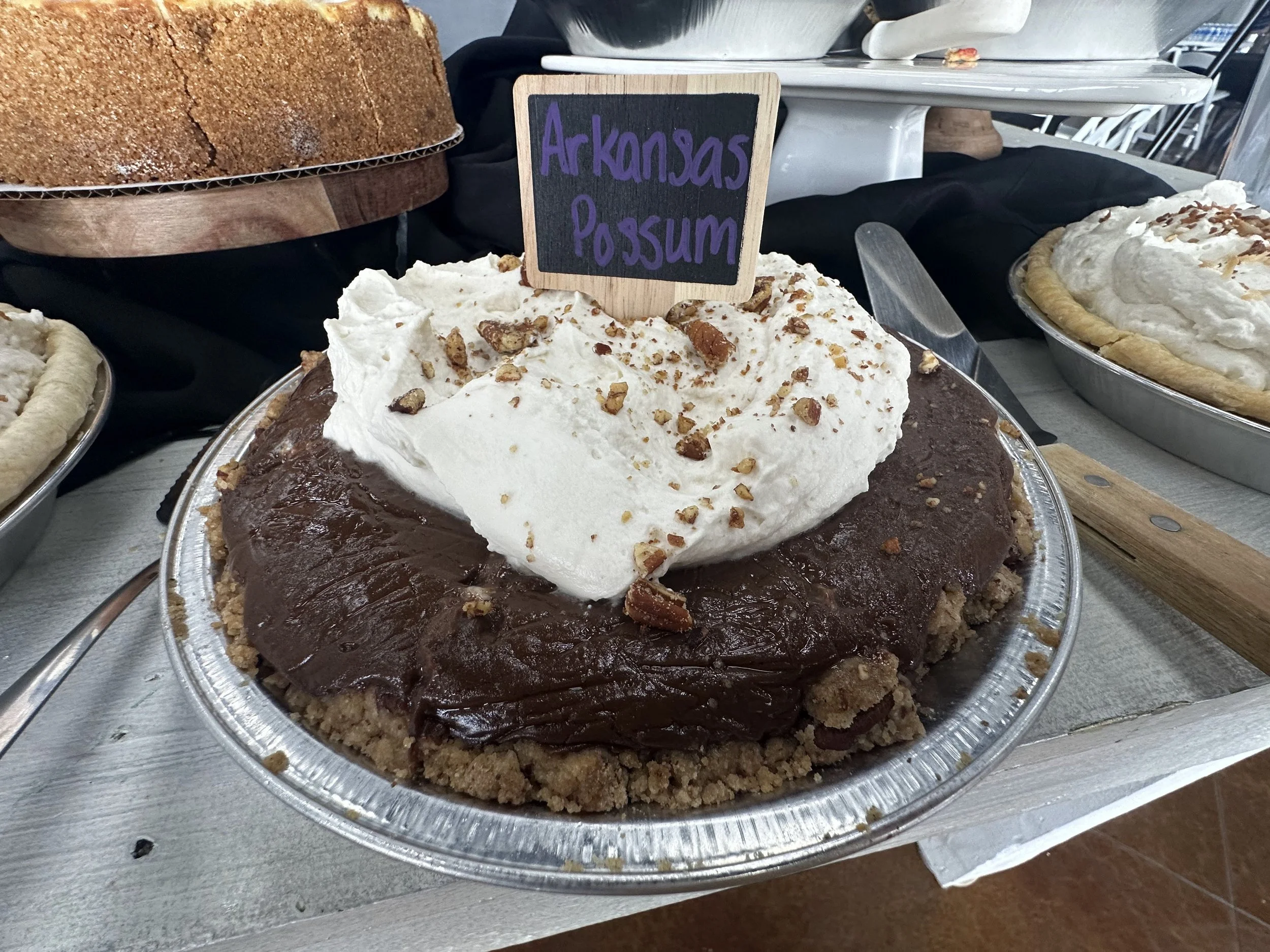 A dessert display featuring a pie labeled 'Arkansas Pecan' and other baked goods, including a chocolate pie with whipped cream and chopped pecans and two other pies behind it.