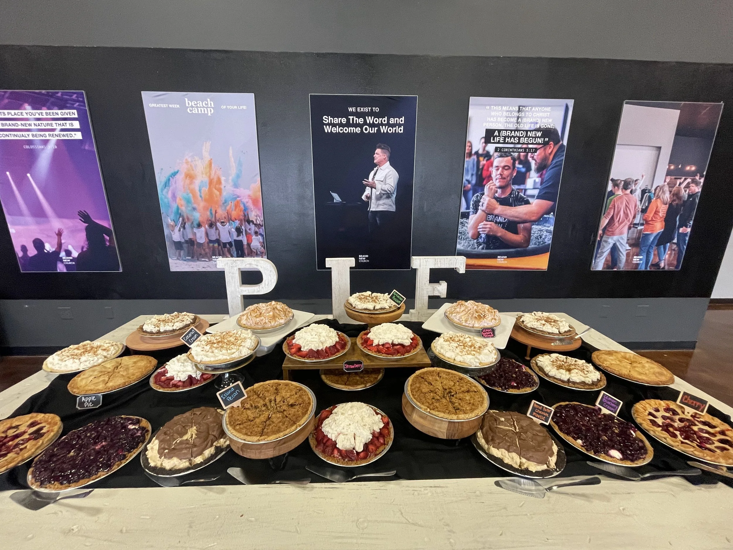 Table of assorted pies and desserts in front of a black wall with posters and photographs. The desserts include apple pie, strawberry pie, island bean, peanut butter chocolate mousse, blueberry pie, and more.