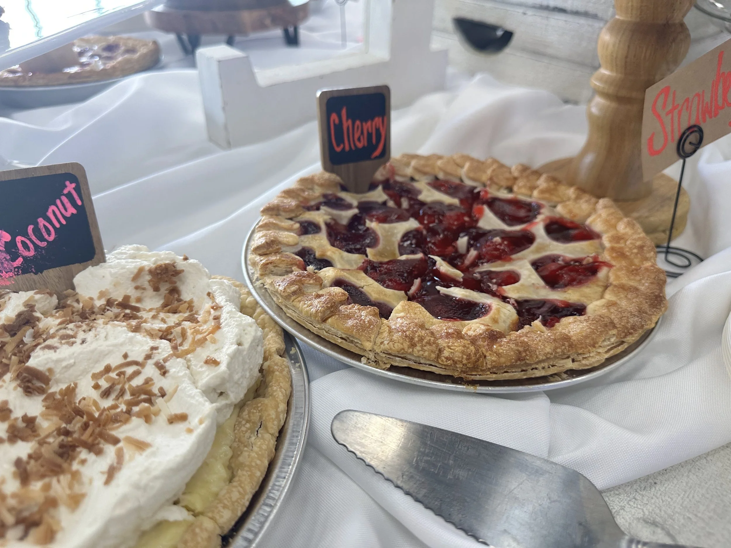 A close-up of two pies on a white table. One pie has a cherry topping with a golden crust, and the other has a cream topping with chopped nuts. Small signs label the pies as 'Cherry' and 'Strawberry' at a bakery or dessert display.