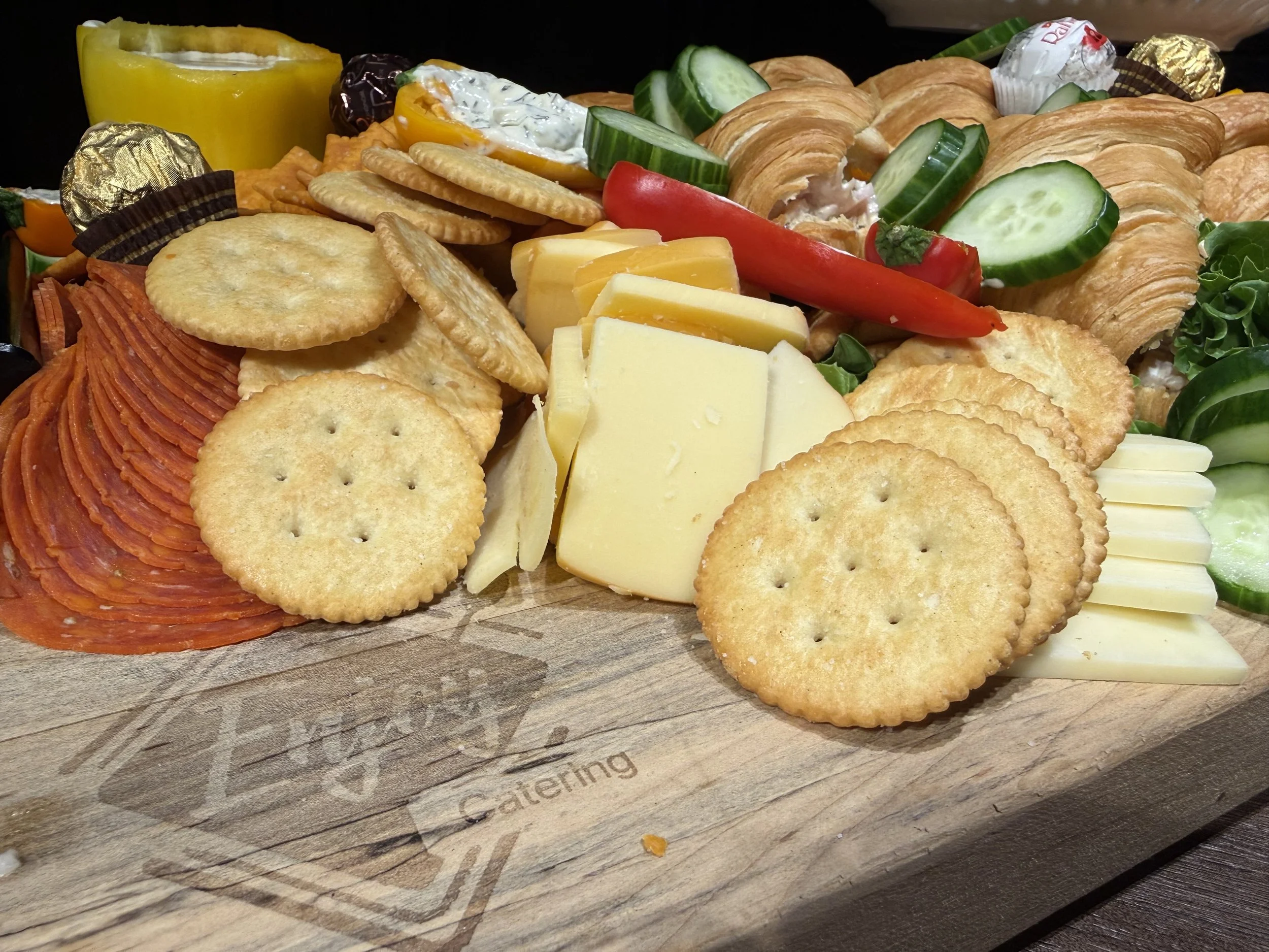 A platter of assorted snacks including crackers, slices of cheese, cucumber, red pepper, yellow pepper, and wrapped chocolates on a wooden surface.