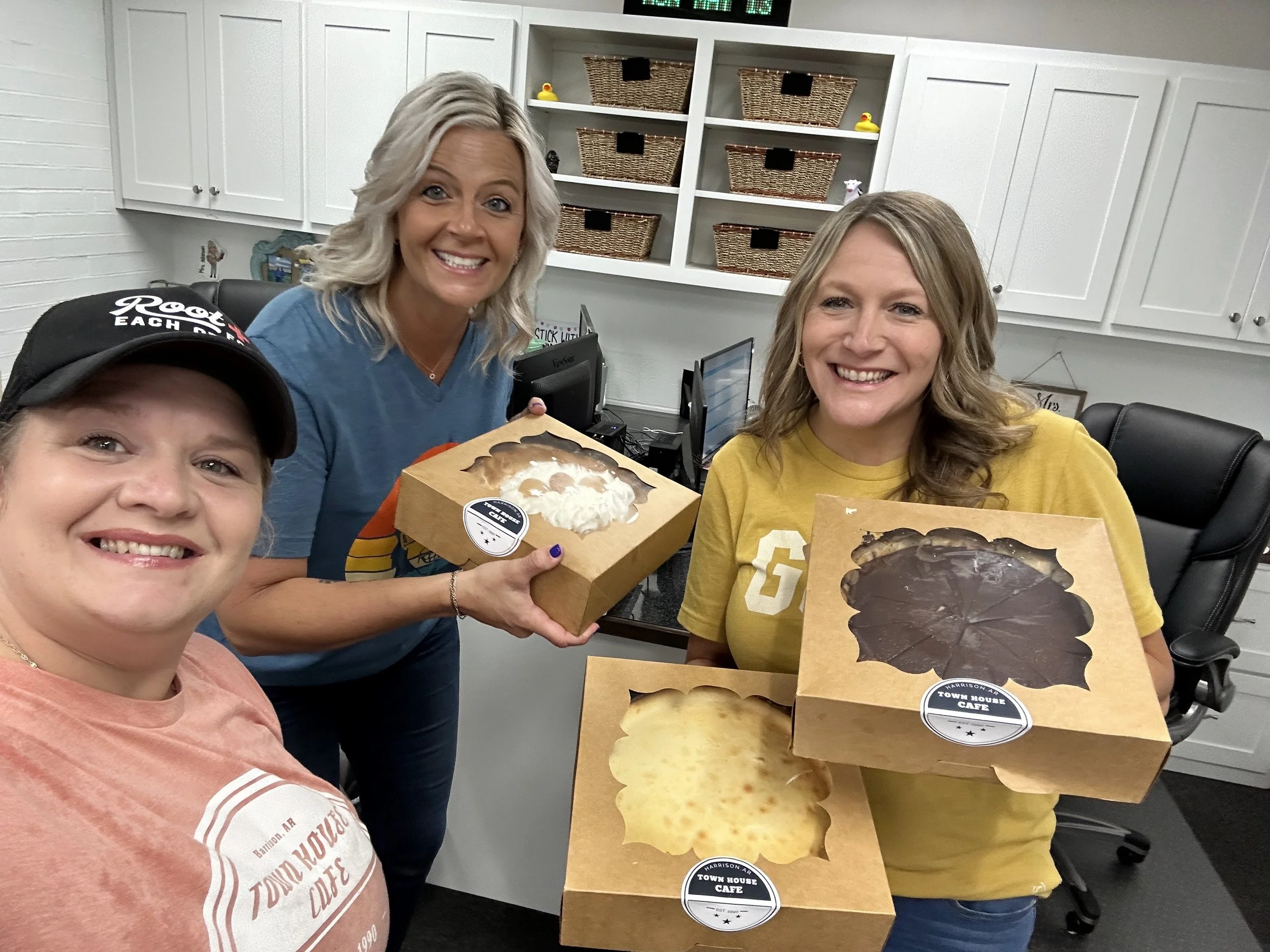 Four women in a room holding boxes of baked goods, with white cabinets and wicker baskets on the wall behind them.