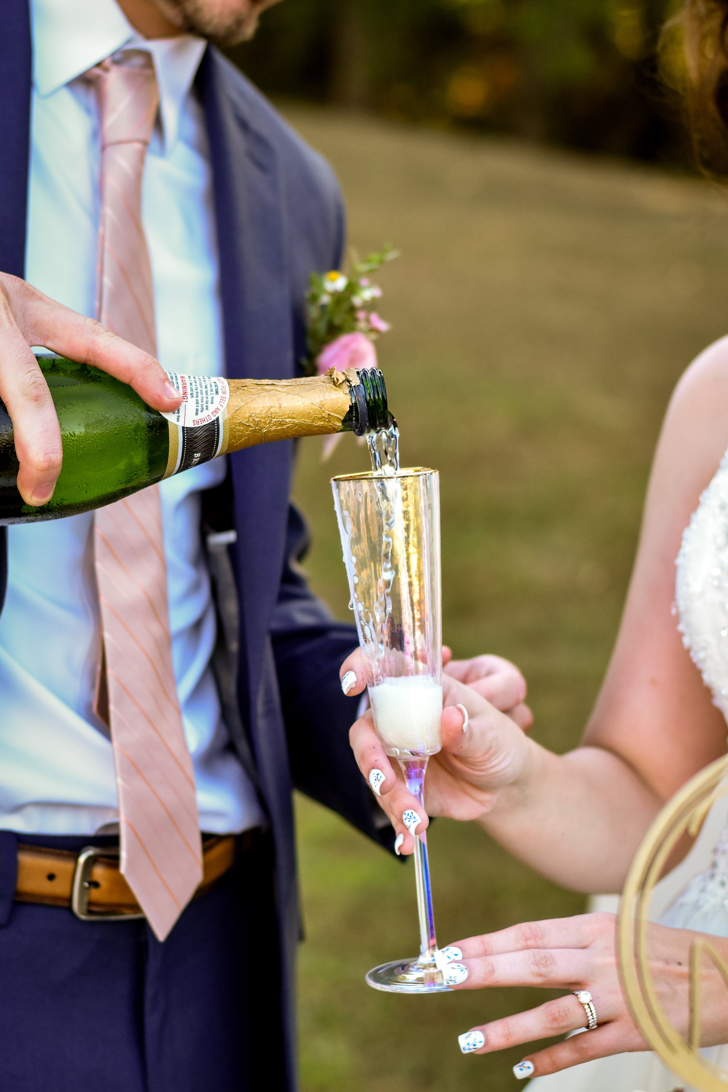 A man in a dark blue suit and a woman in a white dress are at a wedding. The man is pouring champagne into a glass held by the woman, who is wearing a ring and has decorated nails. The woman is dressed in a white wedding gown, and the man has a boutonniere on his suit.